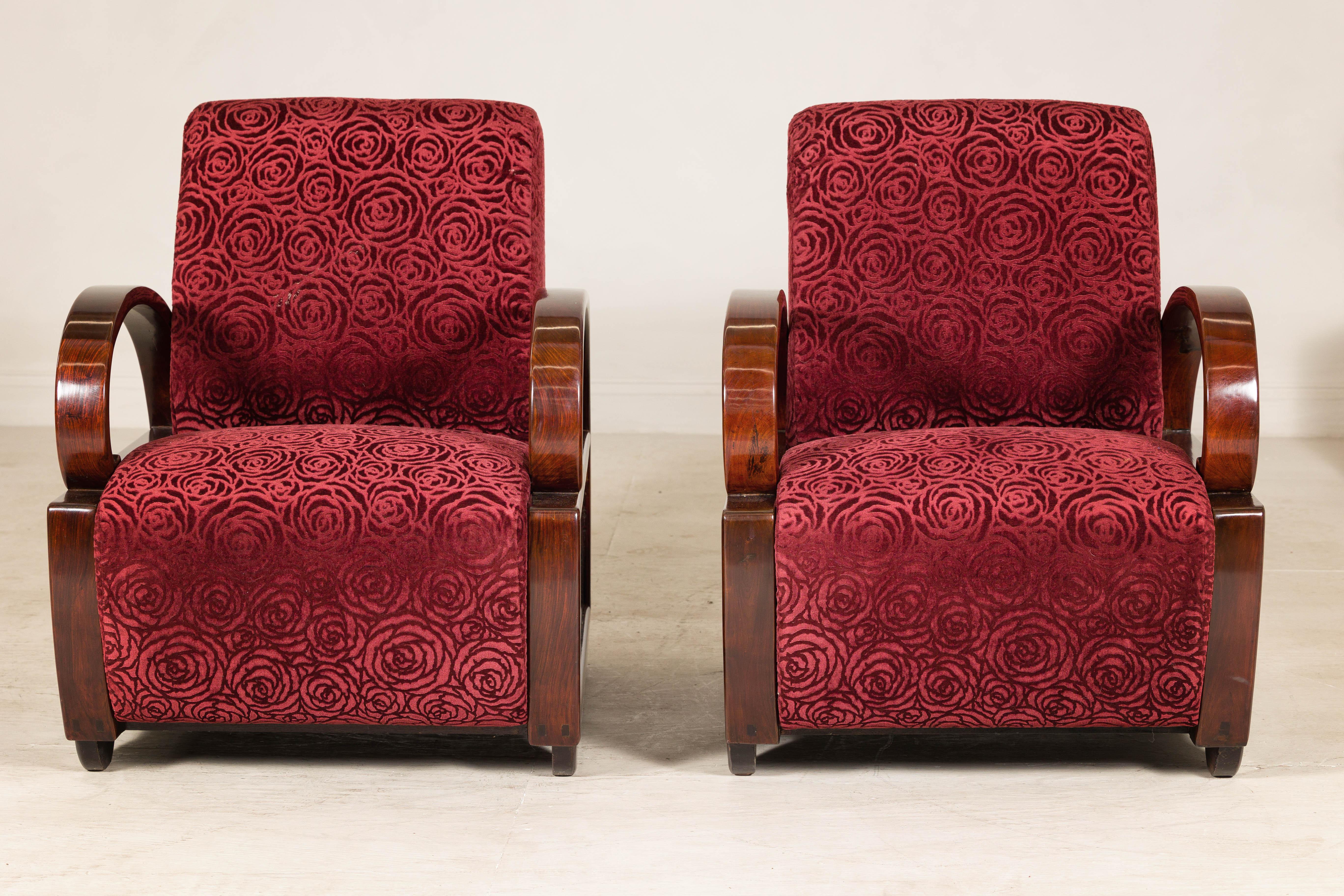 This striking pair of Art Deco club chairs showcases the era's unmistakable blend of bold curves and luxurious materials. Crafted from rich mahogany, the chairs boast gracefully arched arms and sleek side panels, which enhance their sculptural