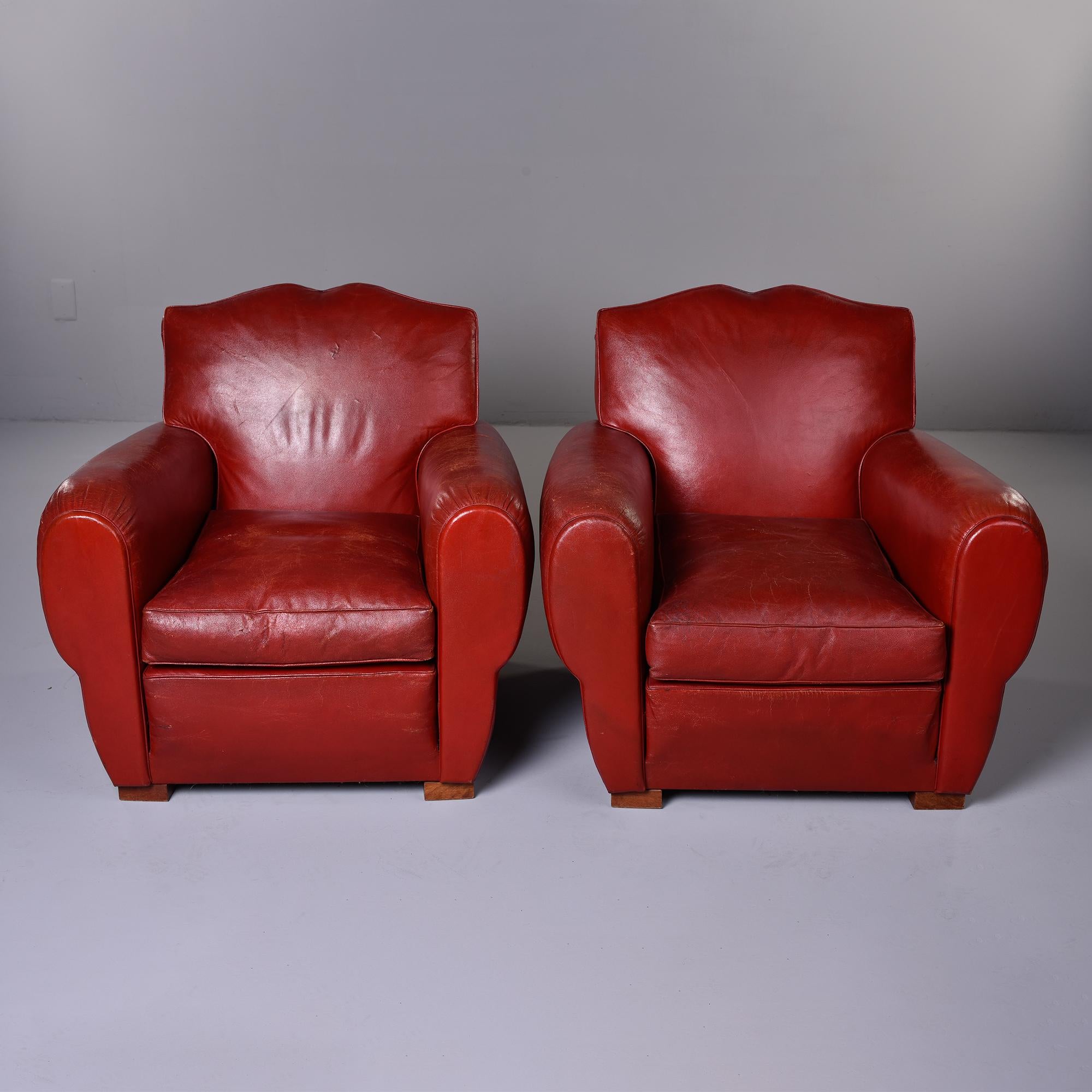 Found in England, this pair of circa 1950s club chairs feature the original deep red leather upholstery and mustache style back rests. Solid construction with spring seats and block feet. Unknown maker. Sold and priced as a pair. Original leather