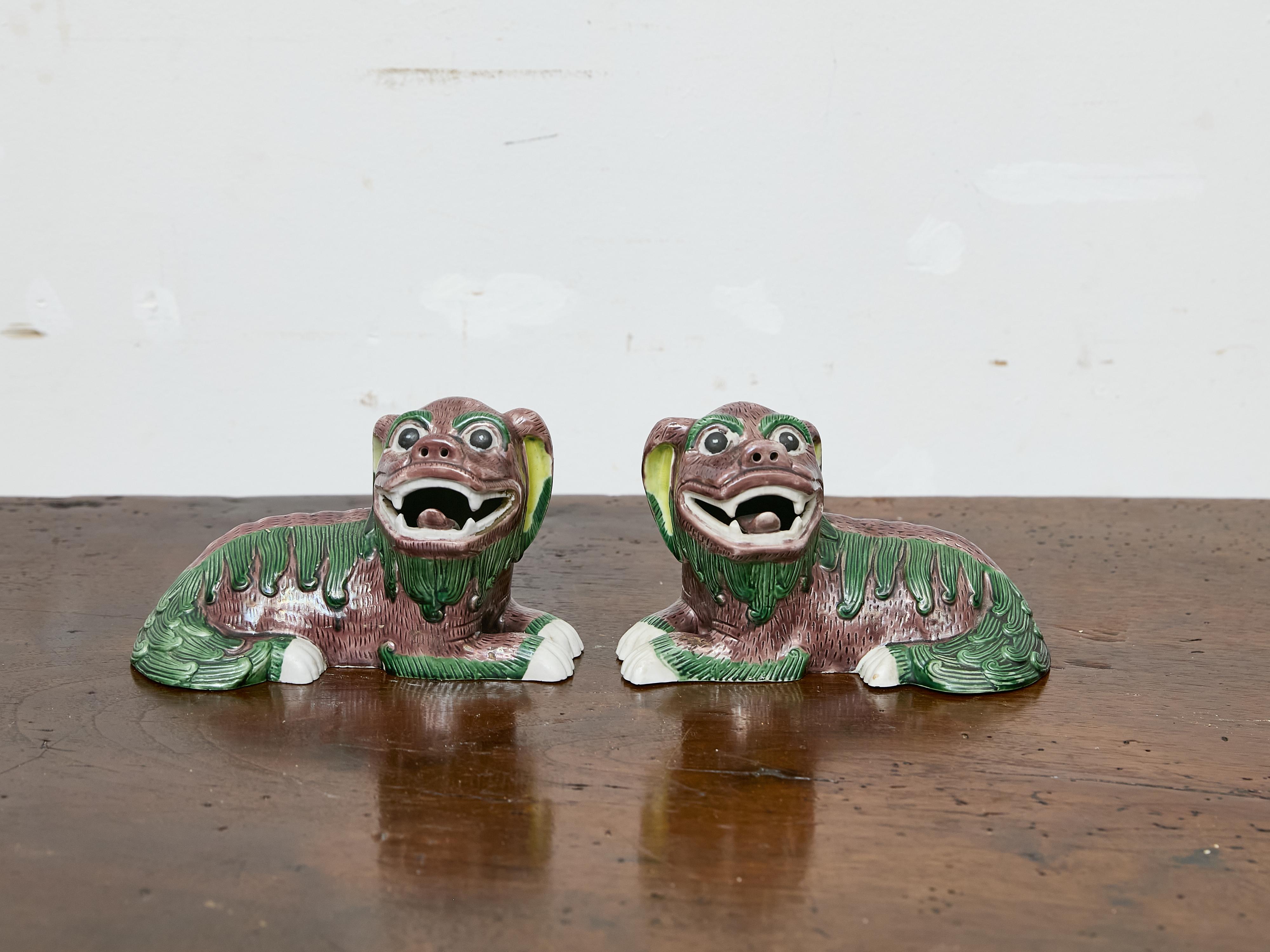 Una stravagante coppia di figure di foo dog in ceramica asiatica vintage, smaltata in una tavolozza di melanzane, verde e bianco. Con le loro espressioni esagerate, le bocche aperte e gli artigli ricurvi, queste figure di guardiani raggiungono un