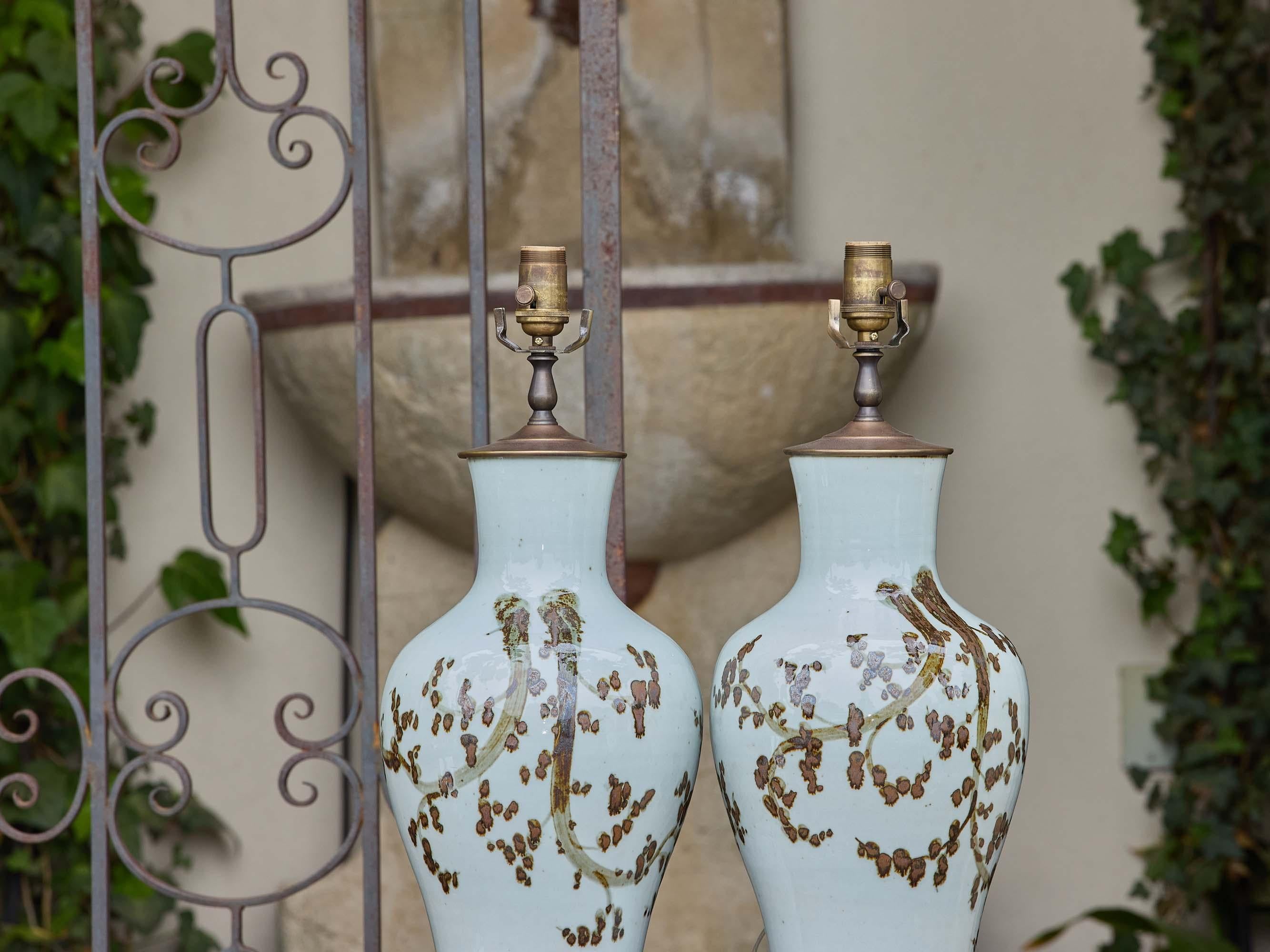 Un par de lámparas de mesa vintage de porcelana de estilo asiático, cada una decorada con un delicado motivo de ramas y flores pintado a mano en ricos tonos marrones y ocres. Los cuerpos redondeados en forma de balaustre están acabados con un