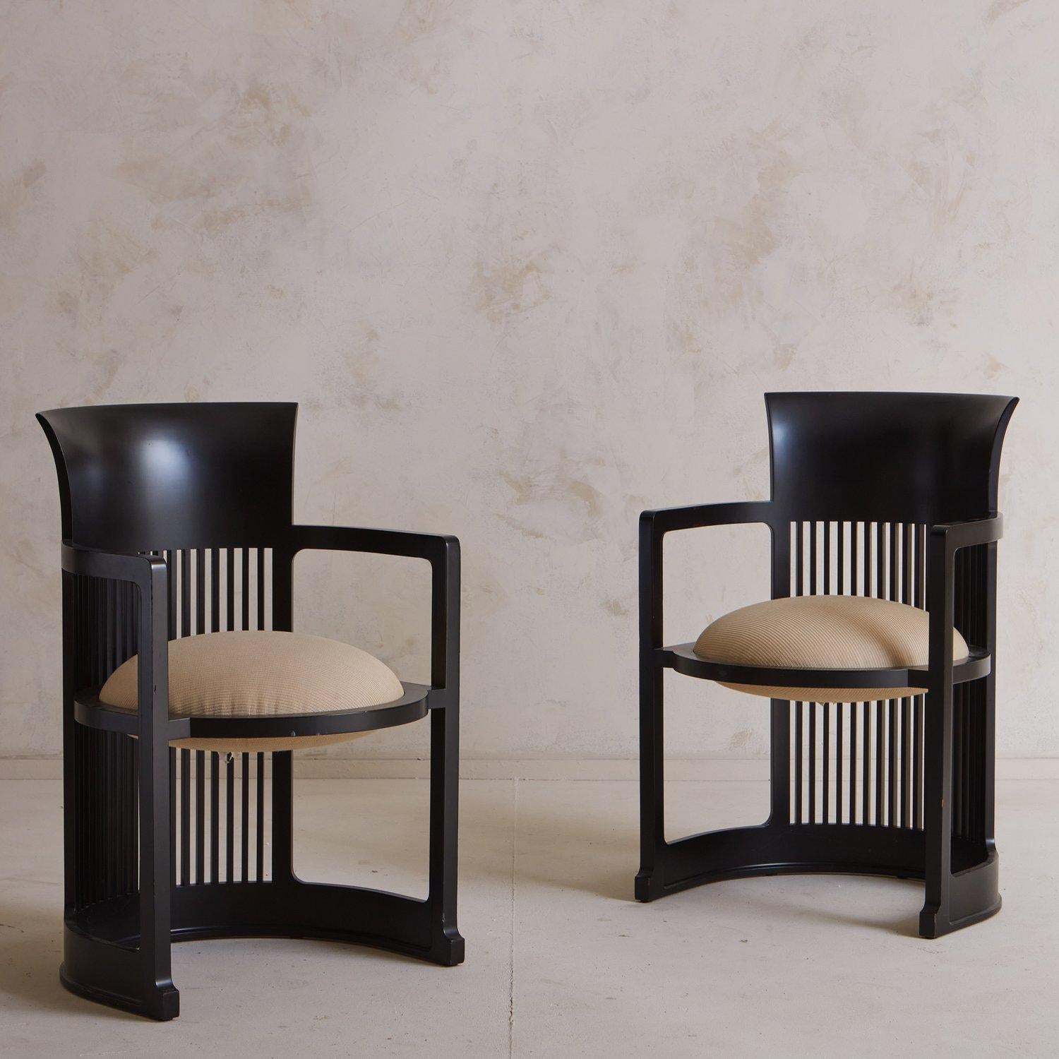 A pair of barrel chairs designed by Frank Lloyd Wright in 1937. These chairs have curved black lacquered wooden frames with a dramatic slatted barrel back. They have angular arms and circular seats upholstered in a textural taupe fabric. Wright