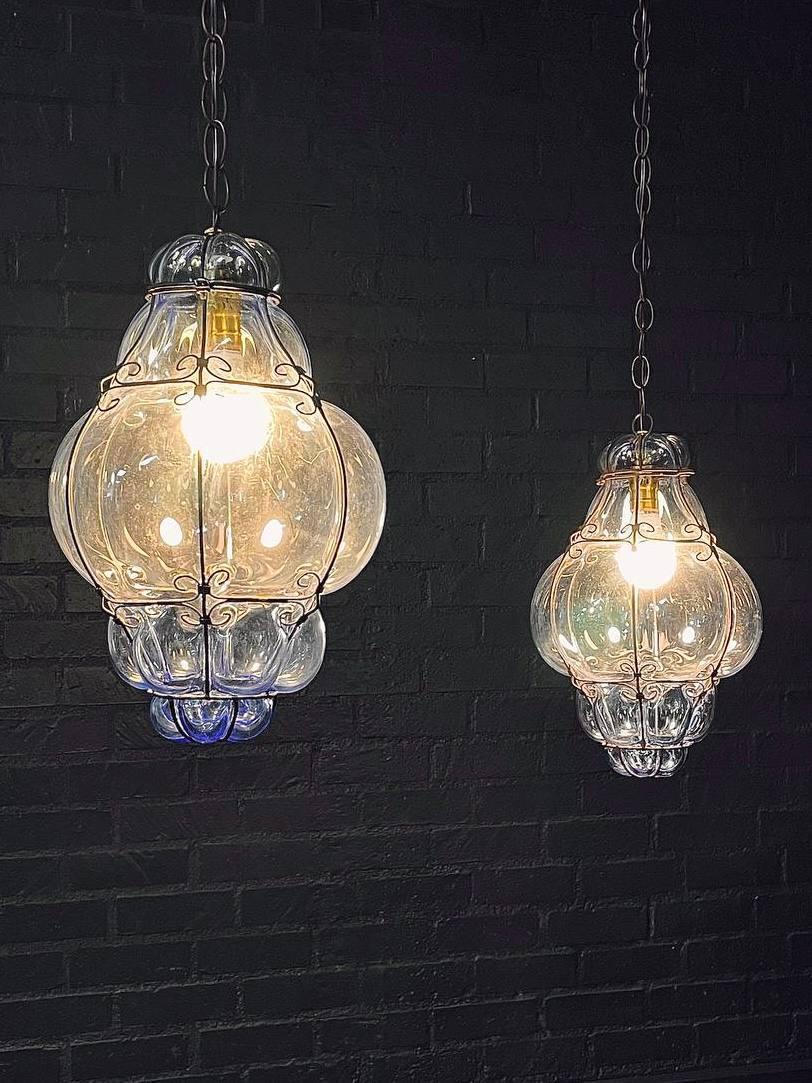 Condition: Newly Rewired 

Materials: Handblown Blue Glass, Wrought Iron Cage, Brass Chain & Canopy 

This stunning pair of vintage pendant lanterns features handblown blue bubble glass encased in ornate wrought iron cages, showcasing the charming