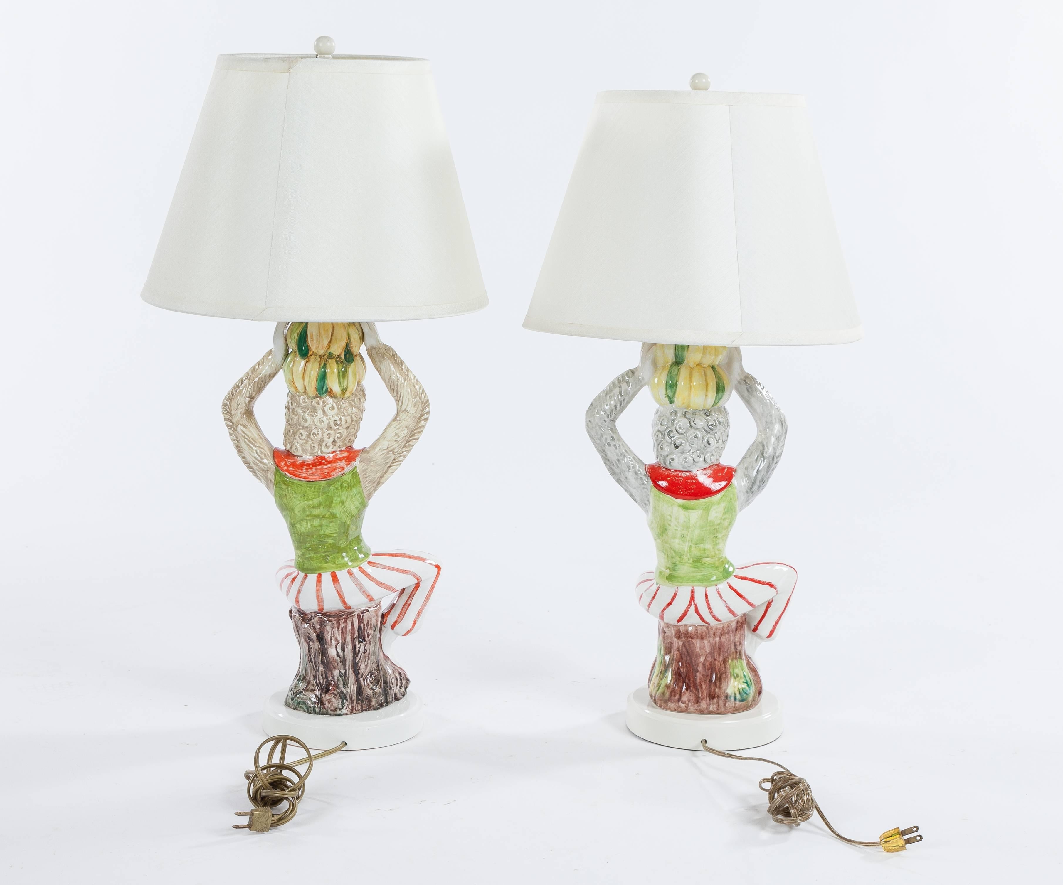 A whimsical pair of vintage ceramic monkey lamps dating to the late 20th century. Featuring cream silk shades and two light bulb sockets. One lamp measures approximately 7