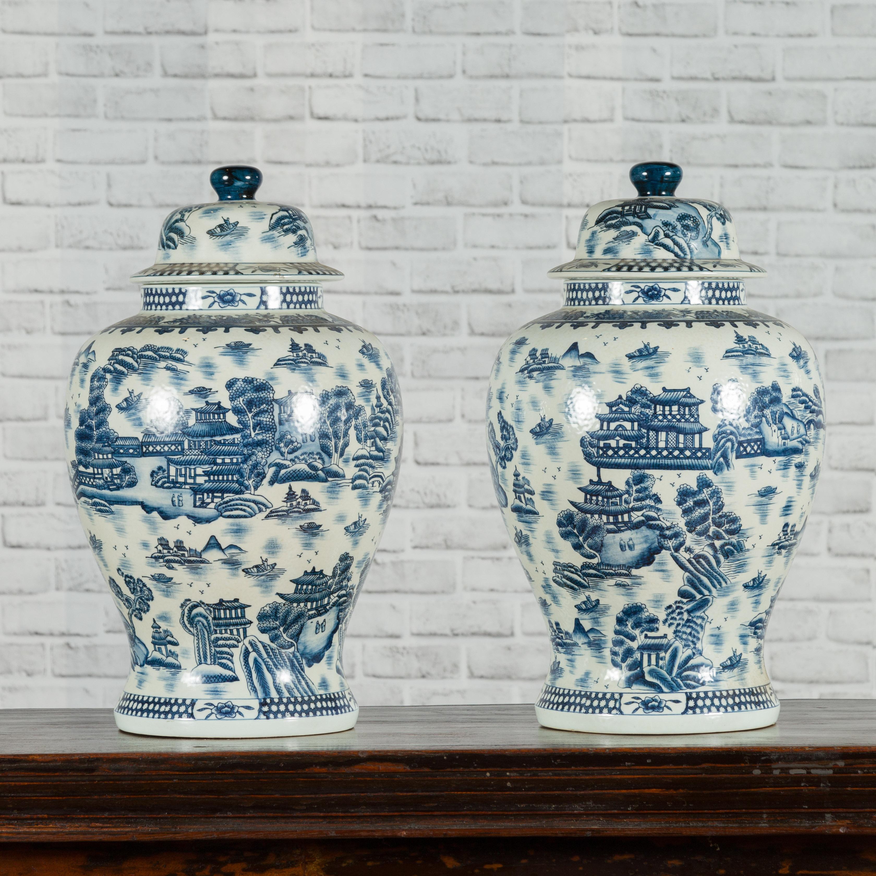 A pair of Chinese vintage blue and white porcelain lidded temple jars from the mid-20th century with landscape and architecture patterns. Created in China during the midcentury period, each of this pair of blue and white porcelain temple jars