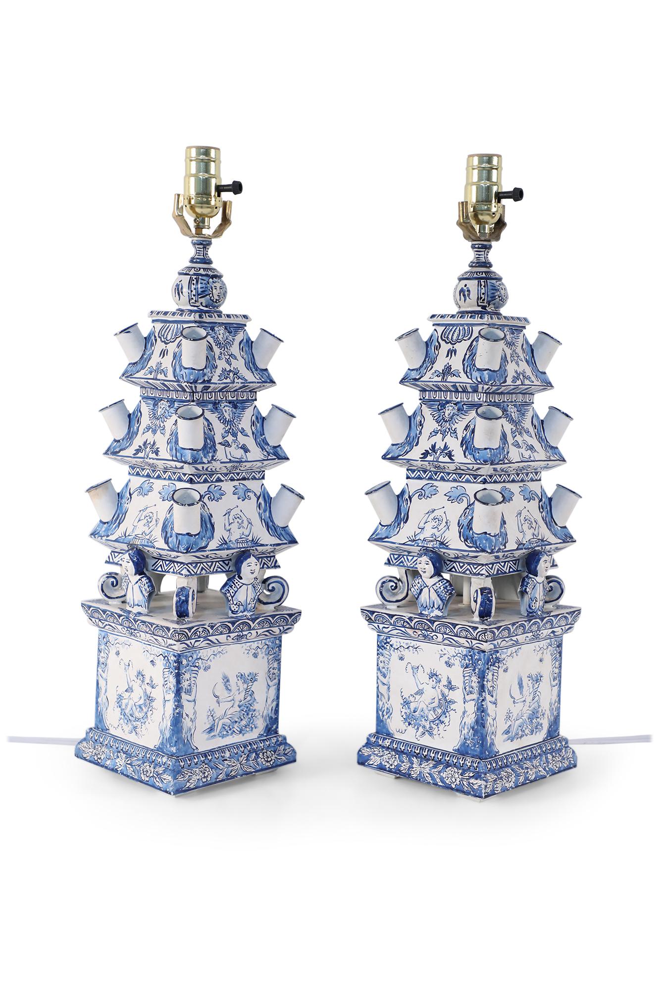 Pair of vintage blue and white tole table lamps created from a tiered tulipiere with designs depicting florals, cherubs, and a dimensional bust of a woman on all four sides, mounted with brass hardware.