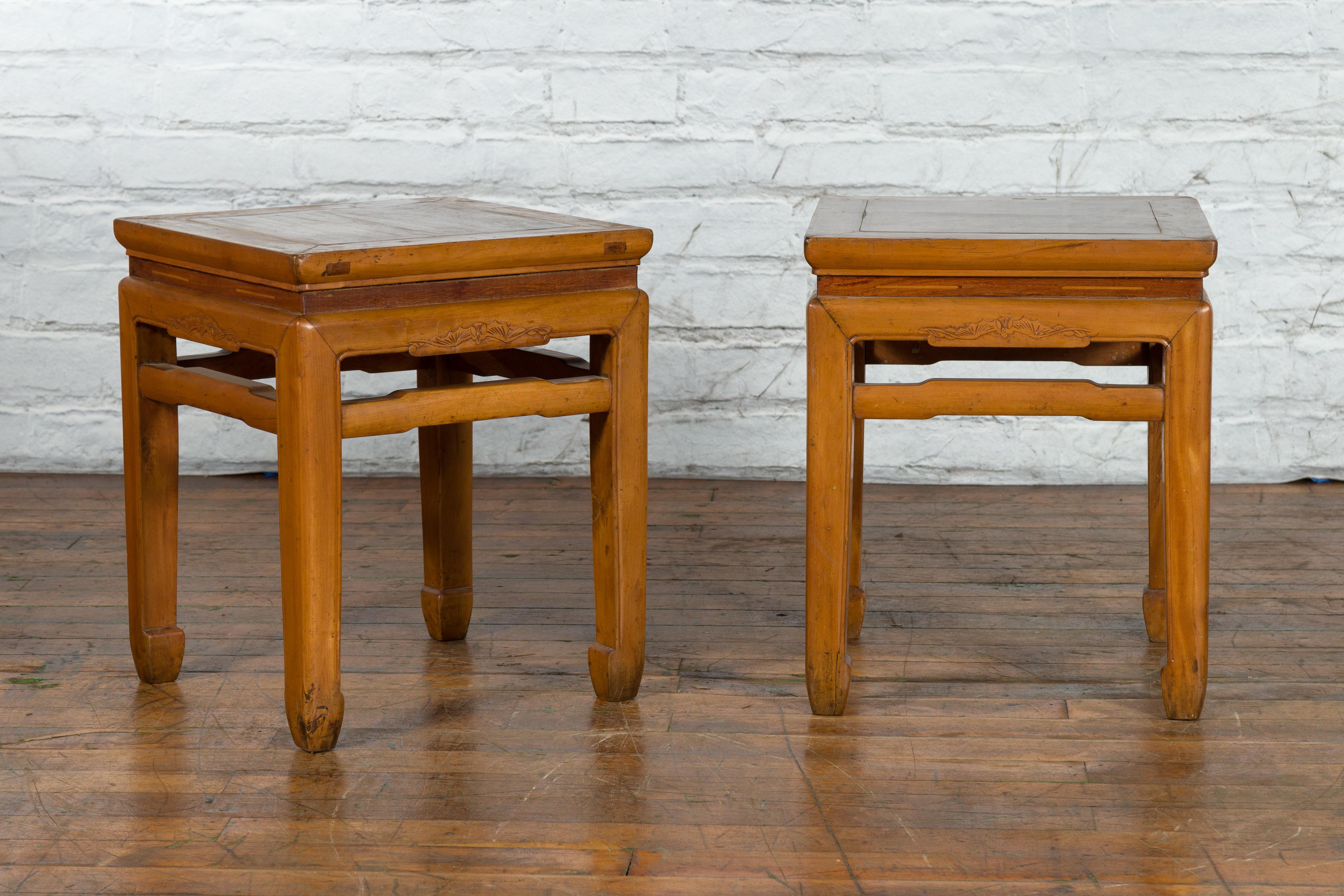 A pair of antique Chinese Ming Dynasty style wooden side tables from the early 20th century, with carved aprons, humpback stretchers and horse hoof legs. Created in China during the Midcentury period, each of this pair of wooden side tables features
