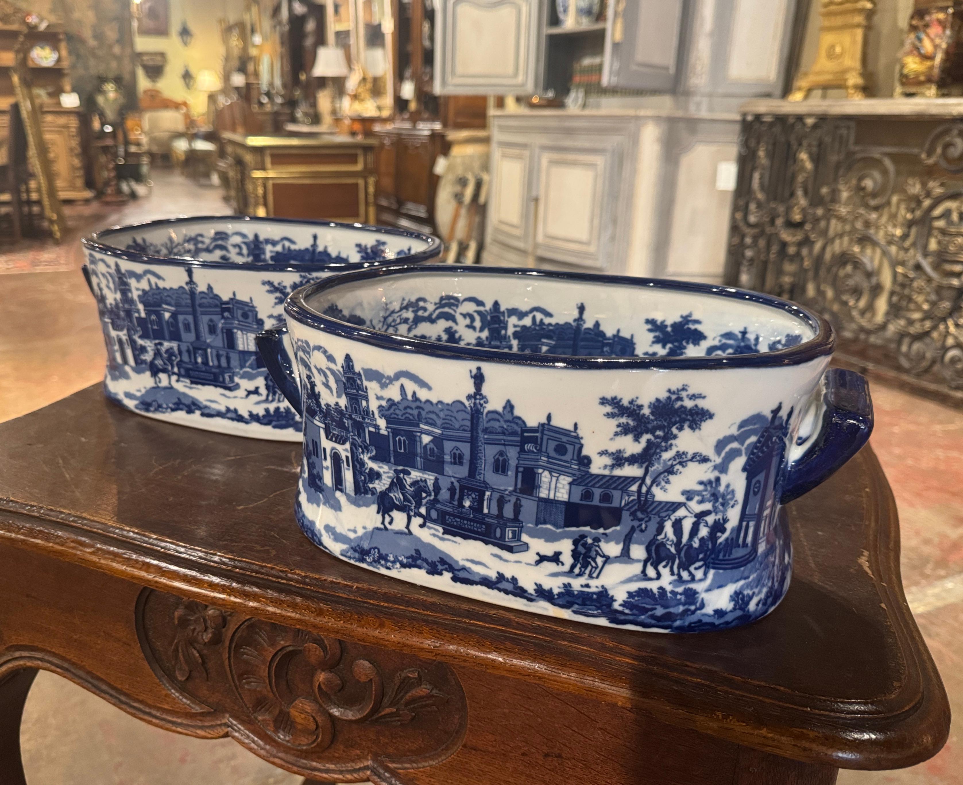 Realizzata in Cina intorno al 1990 e di forma ovale, la coppia di pediluvi in porcellana vintage è dotata di due maniglie laterali. L'opera presenta un'intricata scena architettonica, che comprende un monumento a colonne, edifici imponenti,