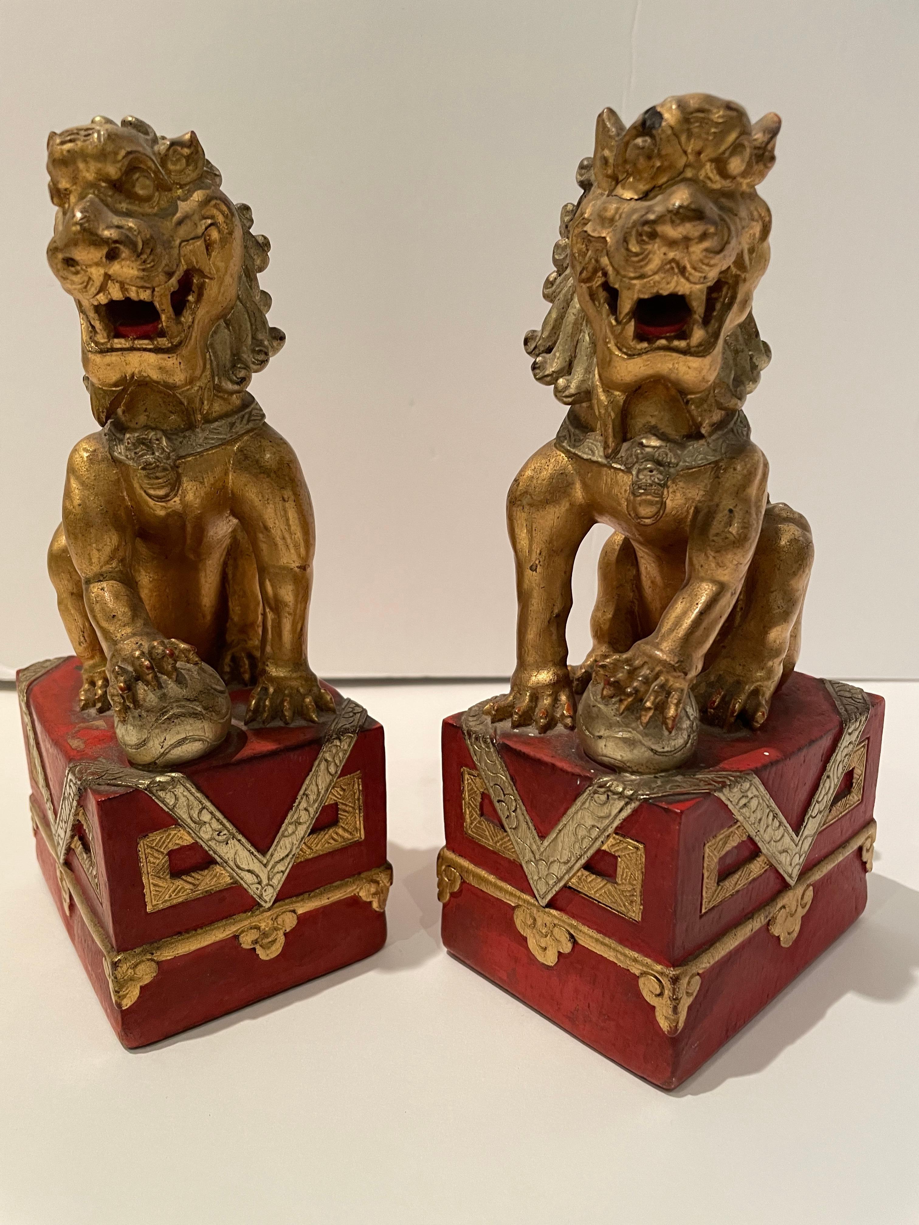 Regal pair of carved wooden Chinese red painted and gilt Buddhistic lions. Each lion is seated atop a cloth draped plinth, with one paw atop a brocade ball. The lion is a symbol of strength and protection. The lion is the protector of the Buddha and