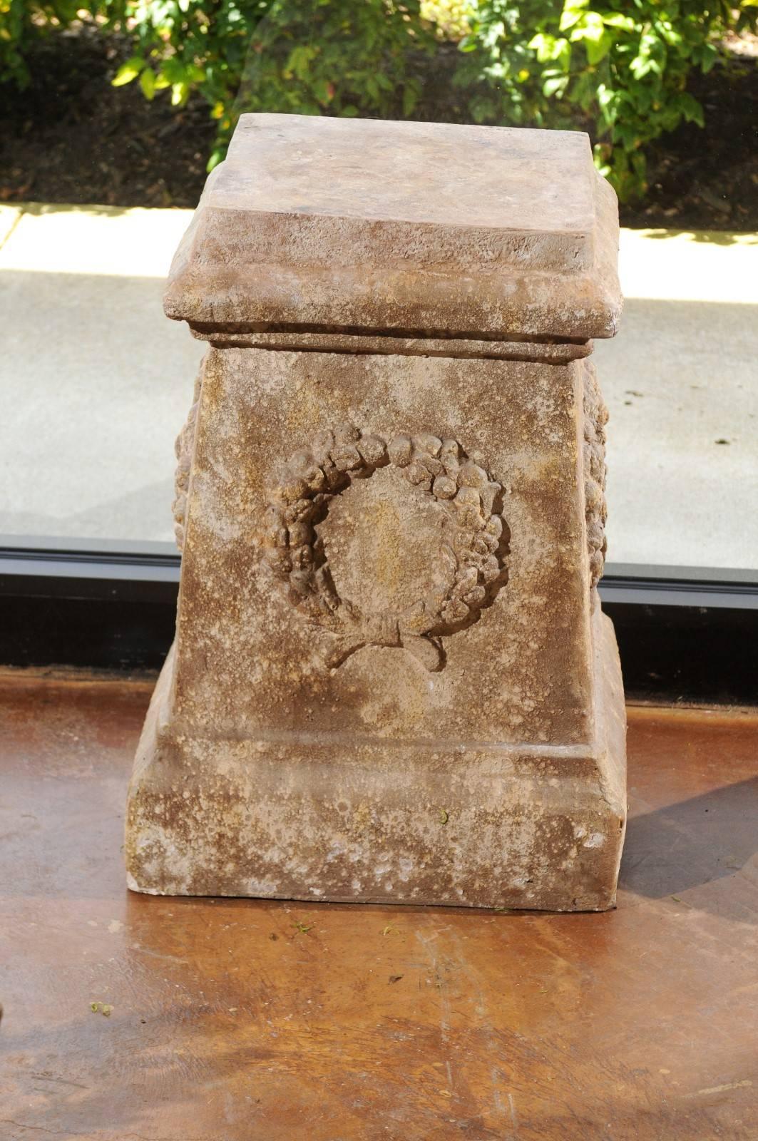 A pair of continental faux stone garden plinths with wreath décor from the mid-20th century. Each of this pair of garden plinths features a raised square top, sitting above a cascading structure. Our eye is immediately drawn to the decorative motifs