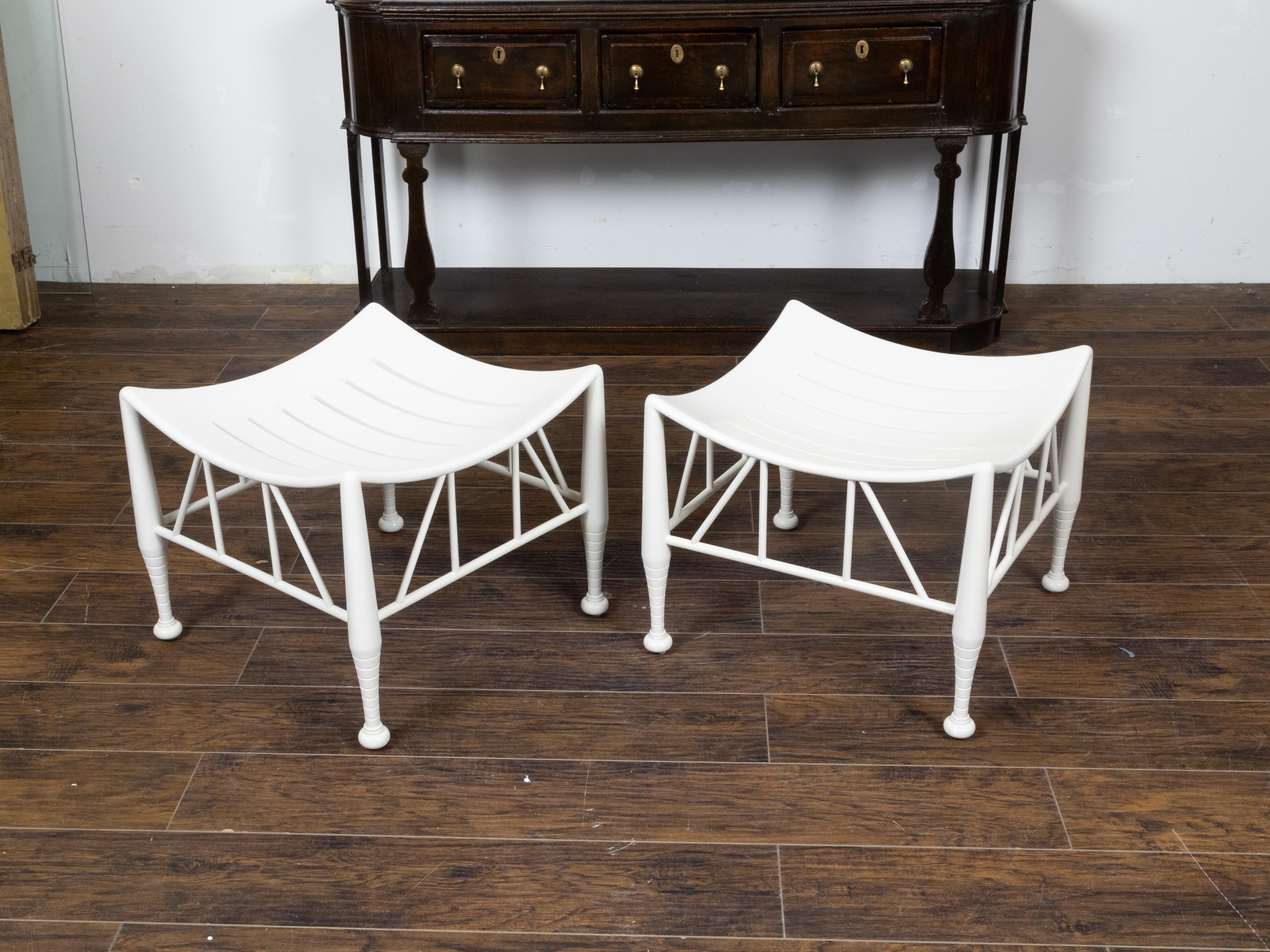 A vintage pair of English Egyptian Revival white painted wood Thebes stool from the mid 20th century, with slatted curving seats, tapering legs on bun feet and intricate aprons. This pair of vintage English Egyptian Revival Thebes stools from the