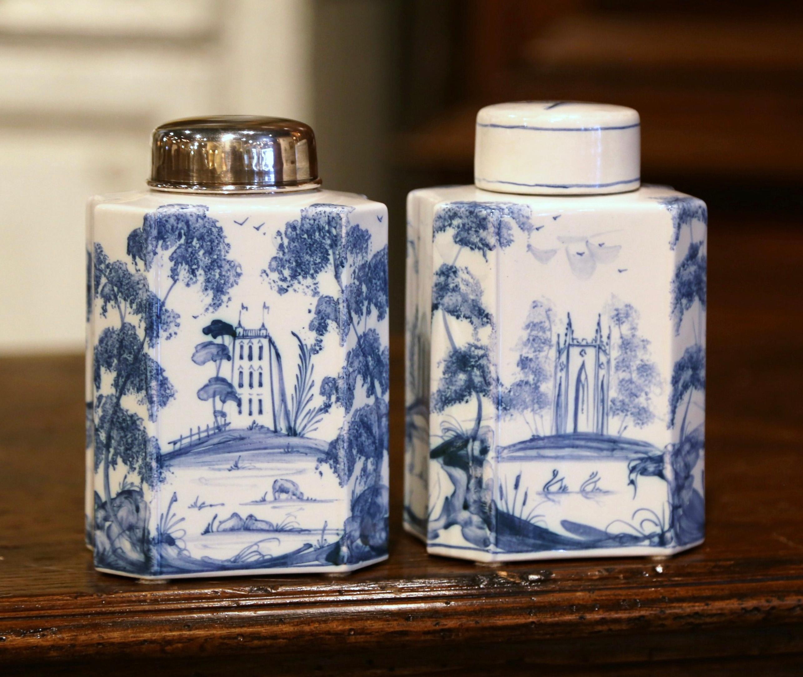 Crafted in Oxford, England circa 1980, each faience jar is hand painted with pastoral motifs in the traditional Delft blue and white palette. One jar has a metal lid, while the other is dressed with a ceramic top. The colorful jars are in excellent