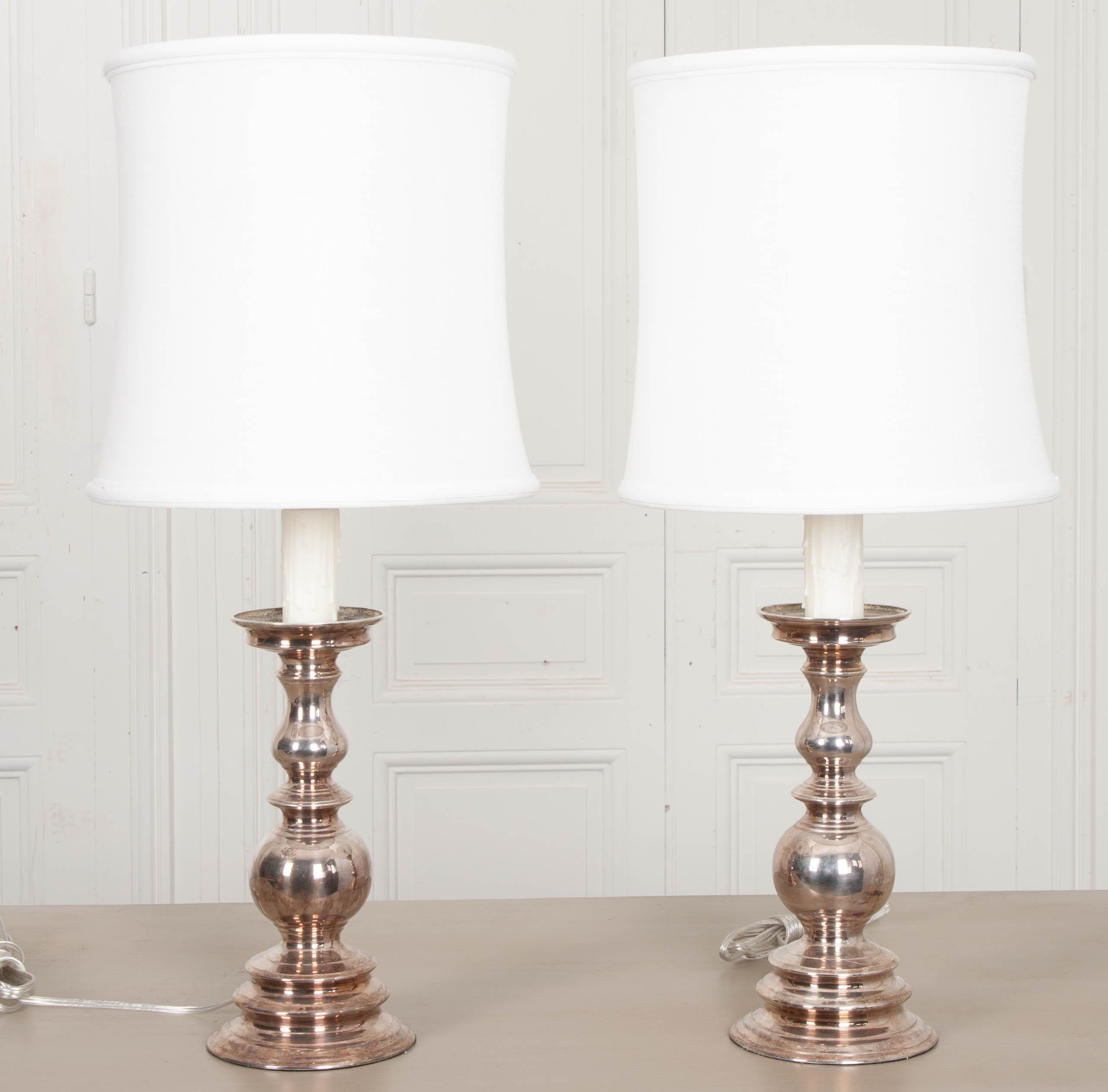 A lovely pair of candlestick-form table lamps, with bases plated in silver. The traditionally-styled duo radiate elegance without a piddling vestige of grandiloquence or pomposity. Included are two white linen, corset-form drum shades. Cleaned and