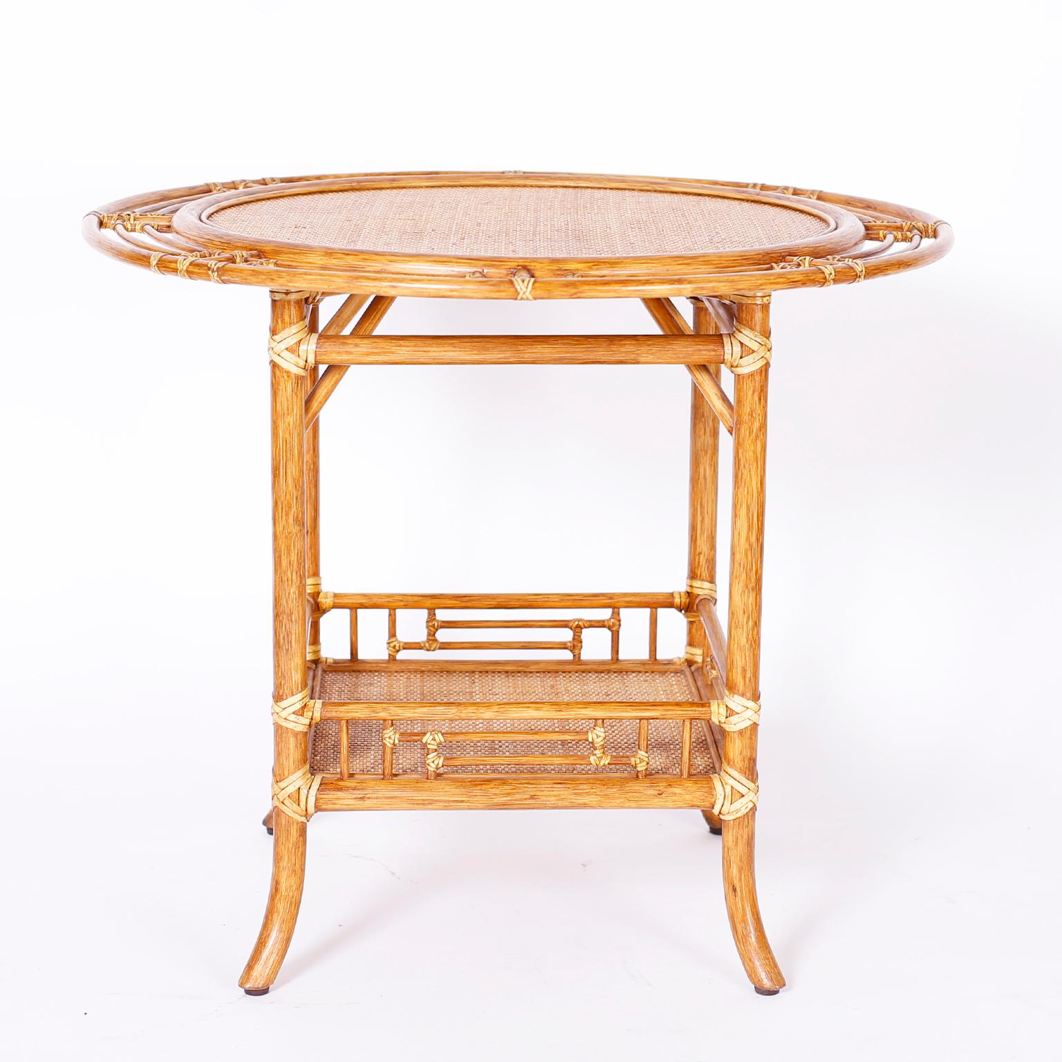 Pair of British colonial style round top two tiered tables crafted with indigenous hardwood frames wrapped with reed and having grasscloth surfaces on both tiers. Labeled MCGuire.