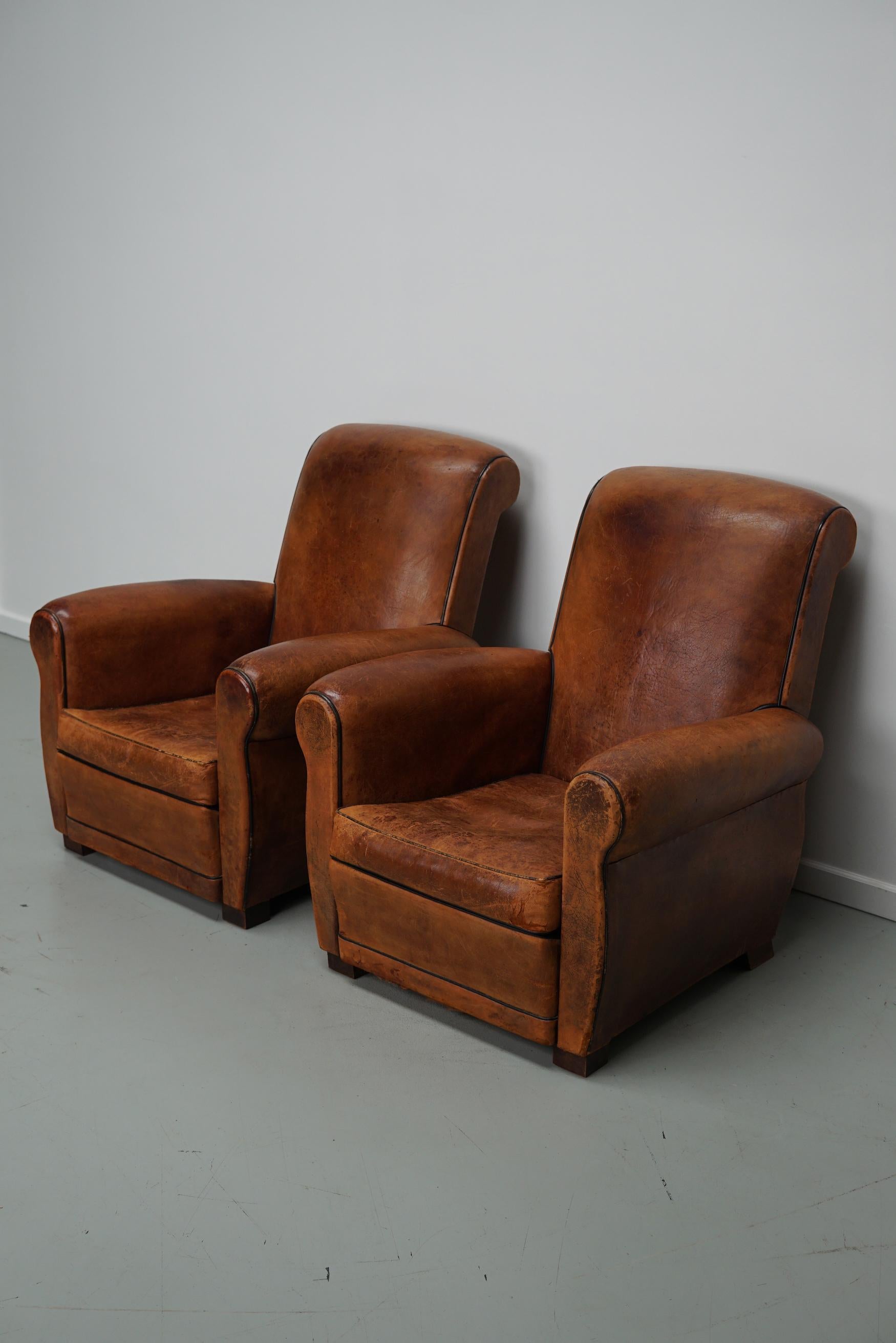 This pair of cognac-colored leather club chairs come from France. They are upholstered with cognac-colored leather and feature metal rivets and wooden legs. They are in a vintage condition and still comfortable.