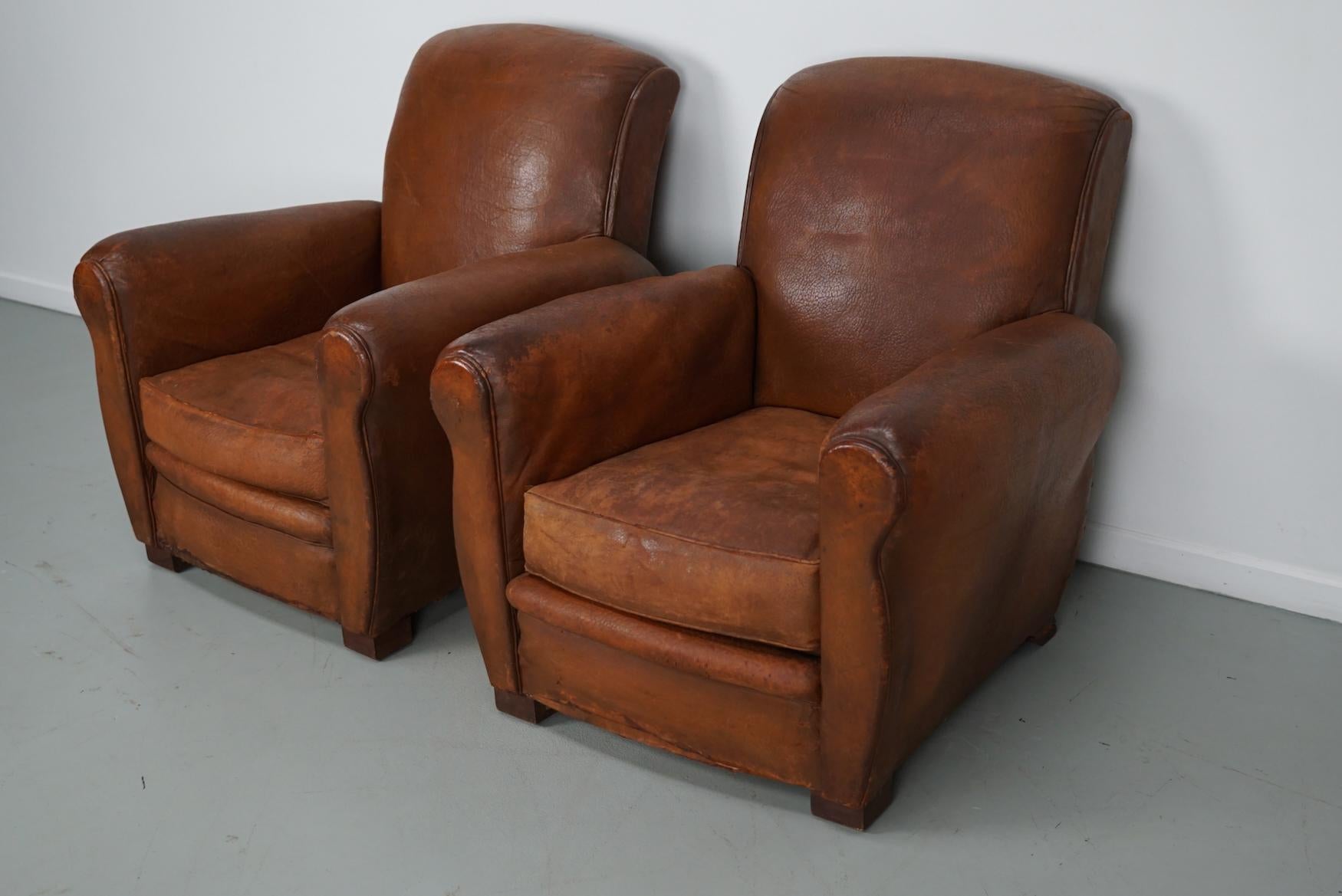 This pair of cognac-colored leather club chairs come from France. They are upholstered with cognac-colored very thick leather and feature metal rivets and wooden legs. They are in a vintage condition and still usable.