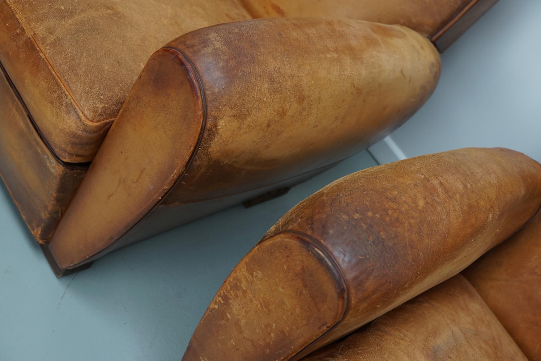This pair of cognac-colored leather club chairs come from France. They are upholstered with cognac-colored leather and feature metal rivets and wooden legs. They are in a vintage condition and still comfortable.