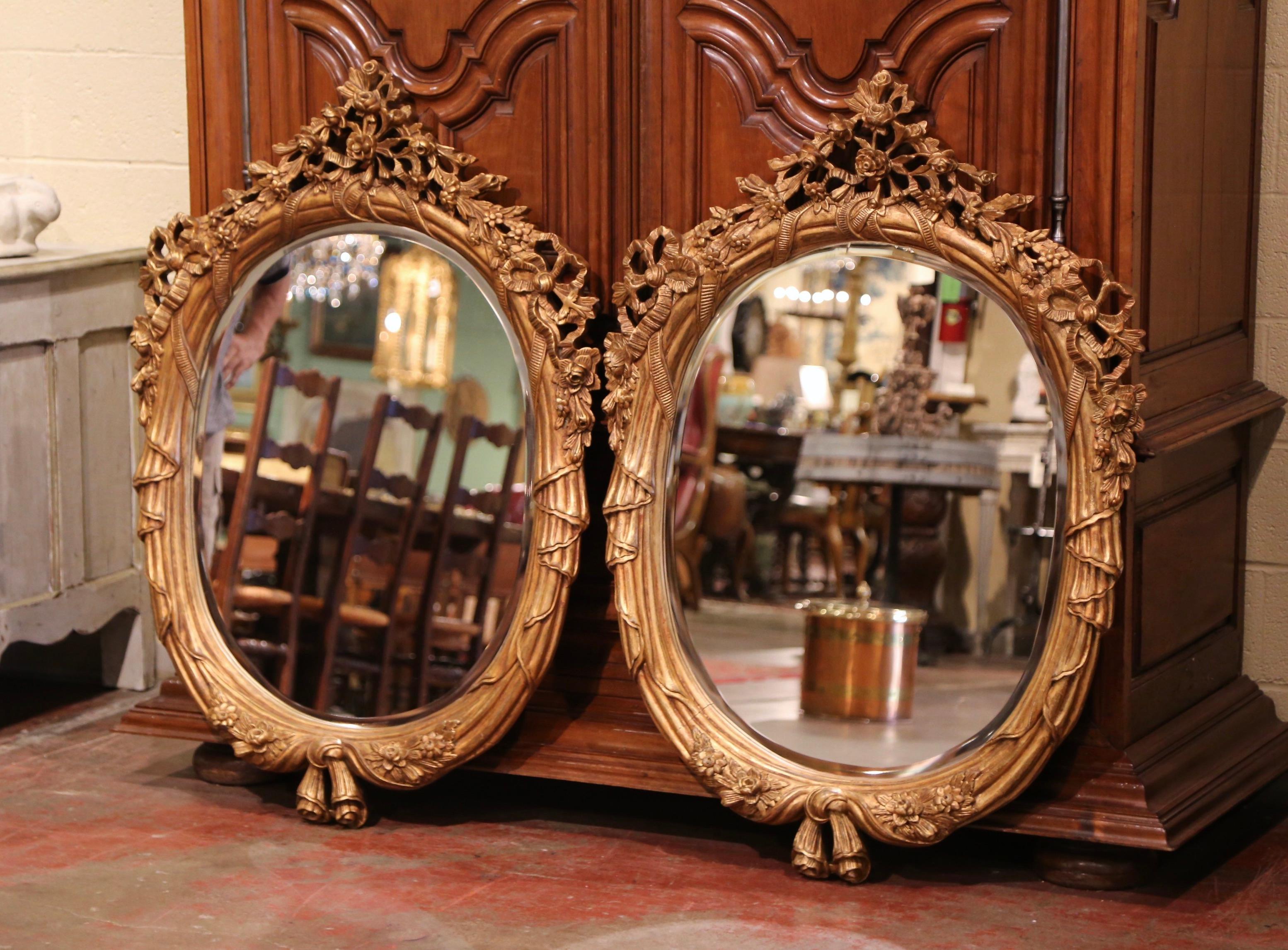 These two matching mirrors were carved in France circa 2000; oval in shape, each mirror is decorated with scrolled and carved motifs at the pediment; the frame is embellished with floral, leaf and tassel motifs tied with a pair of tassels at the