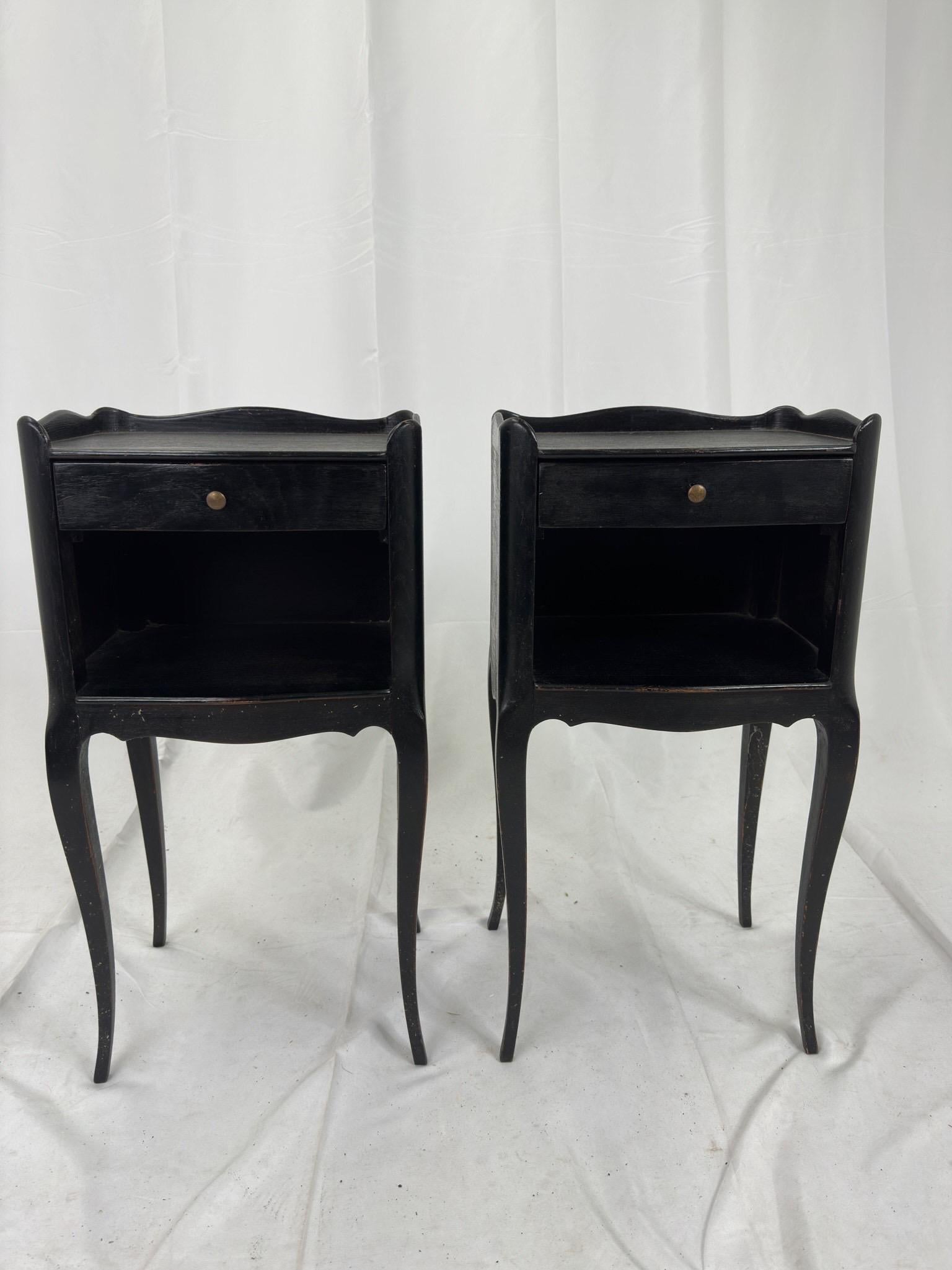 Pairing of vintage French bedside tables in a painted black finish. Each table features a shelf for storage and a single drawer with brass pull. 

Each measure 13.5”W x 10.25”D x 26”H with the top shelf at 25”H. Wear is consistant with age and in