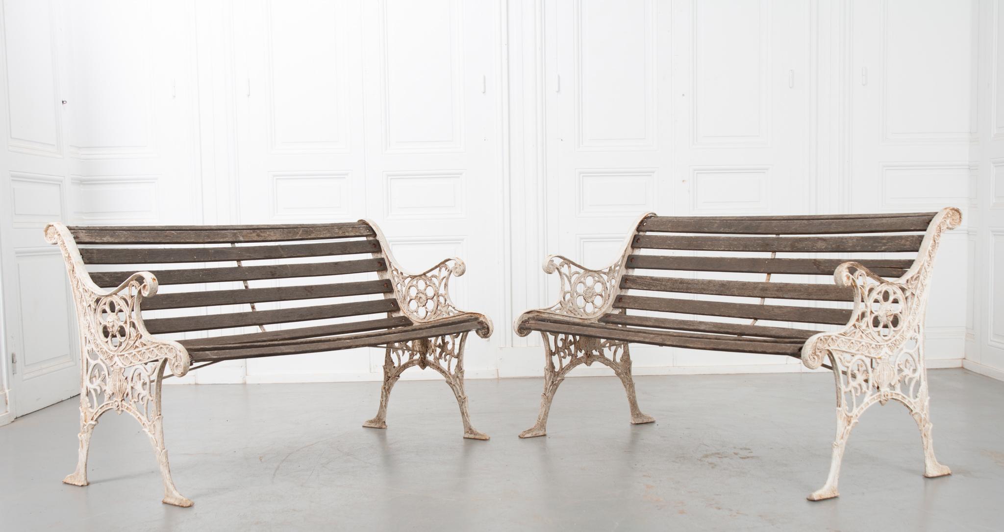 This stunning pair of white painted cast iron benches showcase pierced decorative scroll designs; featuring stars, the face of a lion on the armrest, and a hot air balloon among the leafy motif. The pale worn wood slats create a comfortable seat for