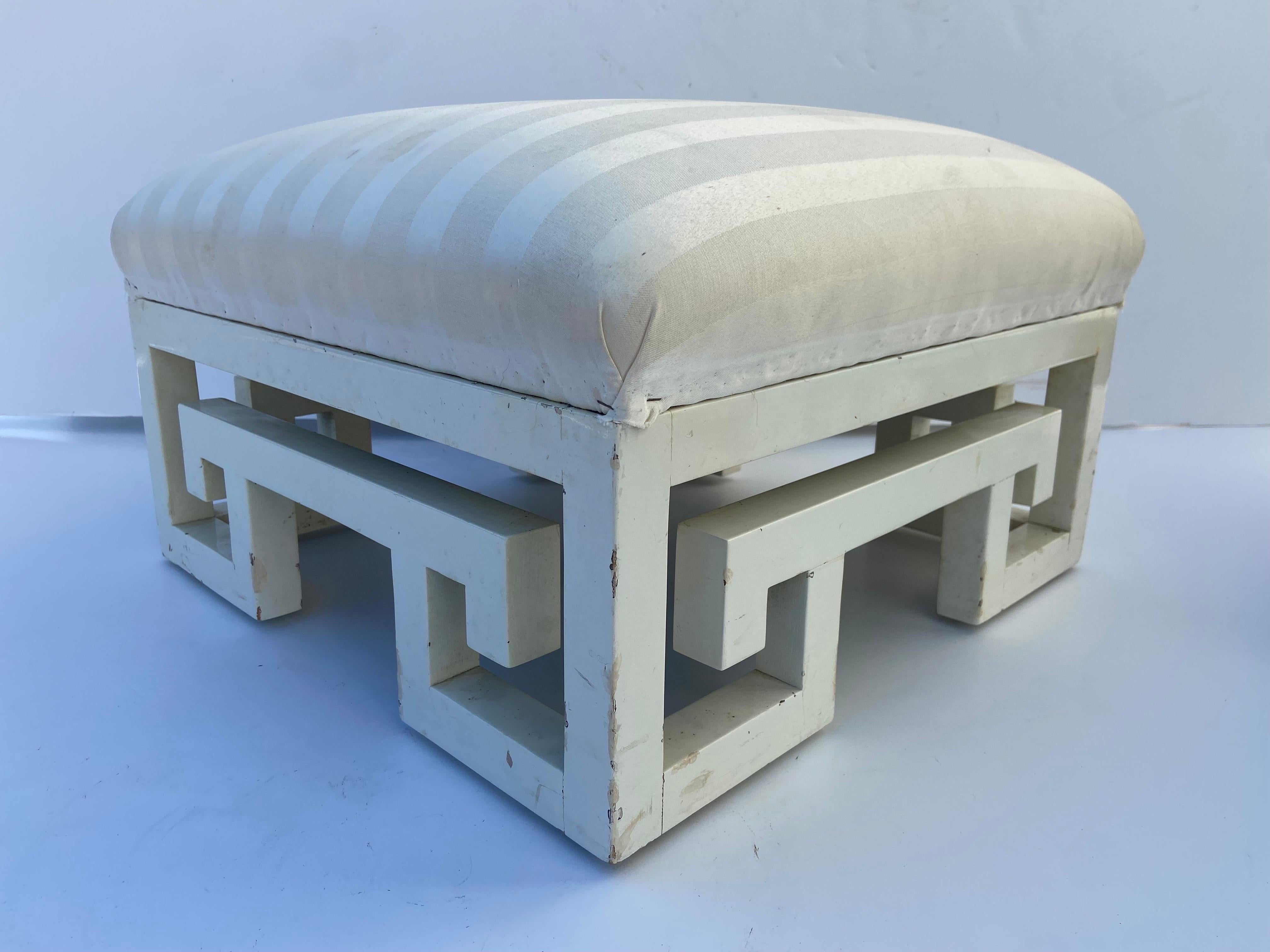 A vintage, circa 1980's, pair of white lacquer painted over wood Greek Key design base ottomans or footstools with upholstery. This upholstered pair is in good vintage condition. The base height is approximately 9 inches without the upholstery. For