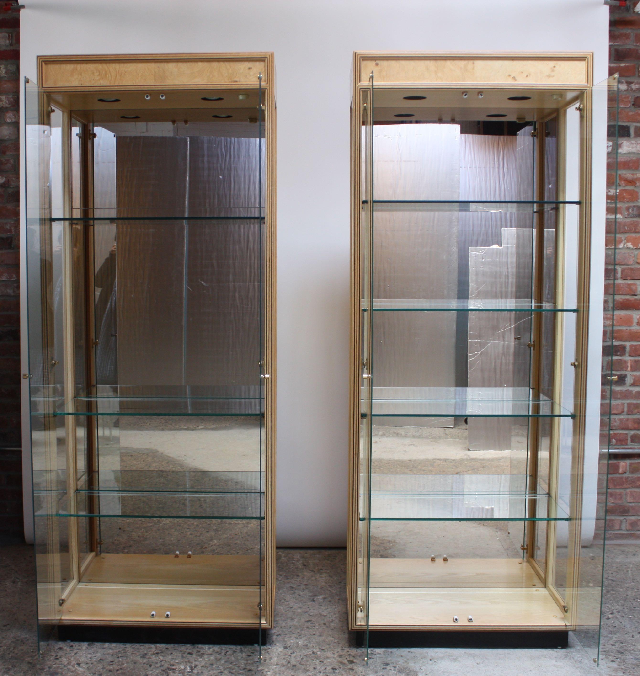 Pair of elegant 1980s Henredon illuminated curio cabinets / display cases beautifully veneered in pale olive burl on ebonized plinth bases.
Details include inlaid sculpted wood trim (which mirrors the ebony of the bases), solid brass hardware,
