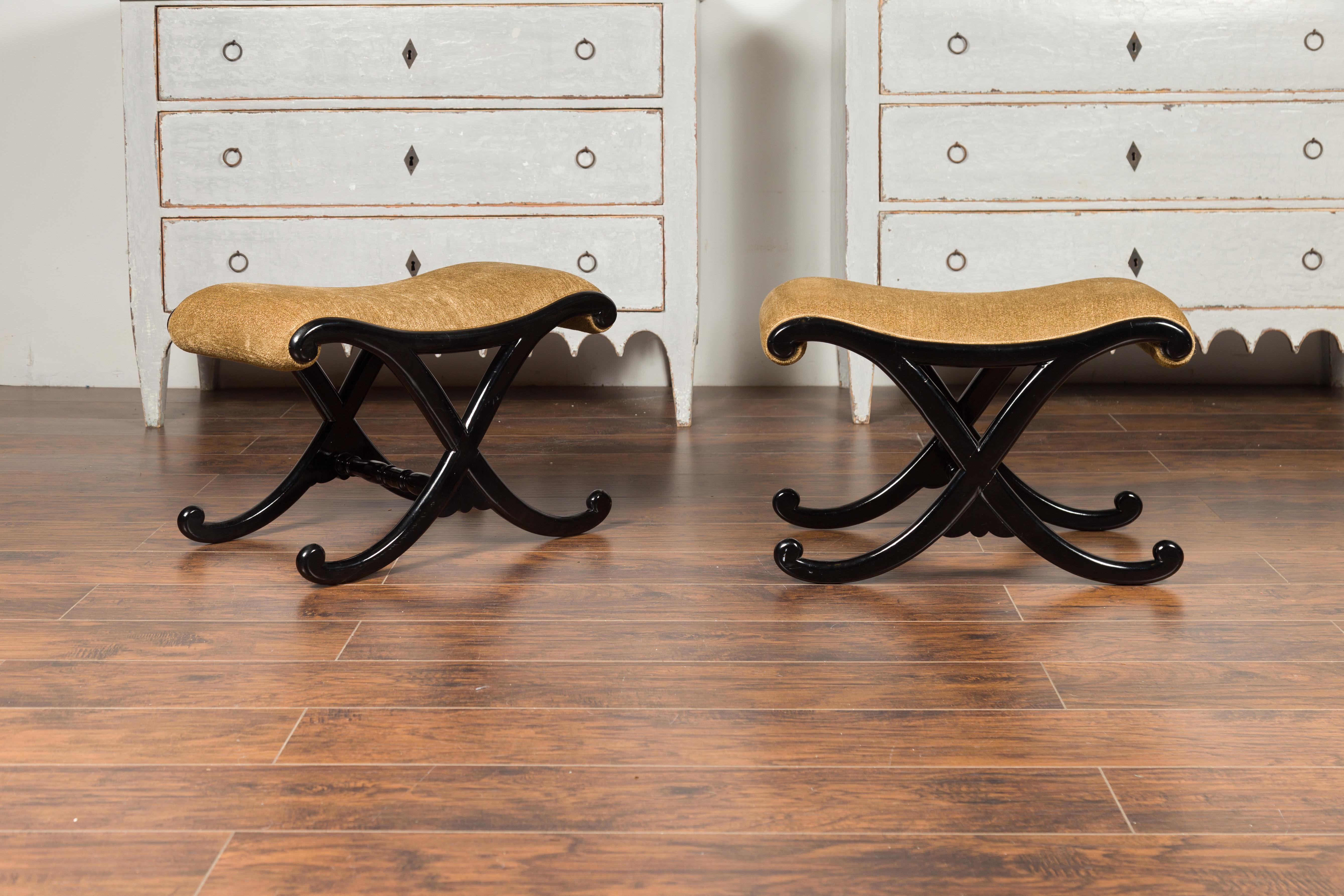 A pair of Italian vintage ebonized wood stools from the mid-20th century, with X-form base and upholstered seats. Born in Italy during the mid-century period, each of this pair of stools features a curving top covered with a tan colored upholstery,