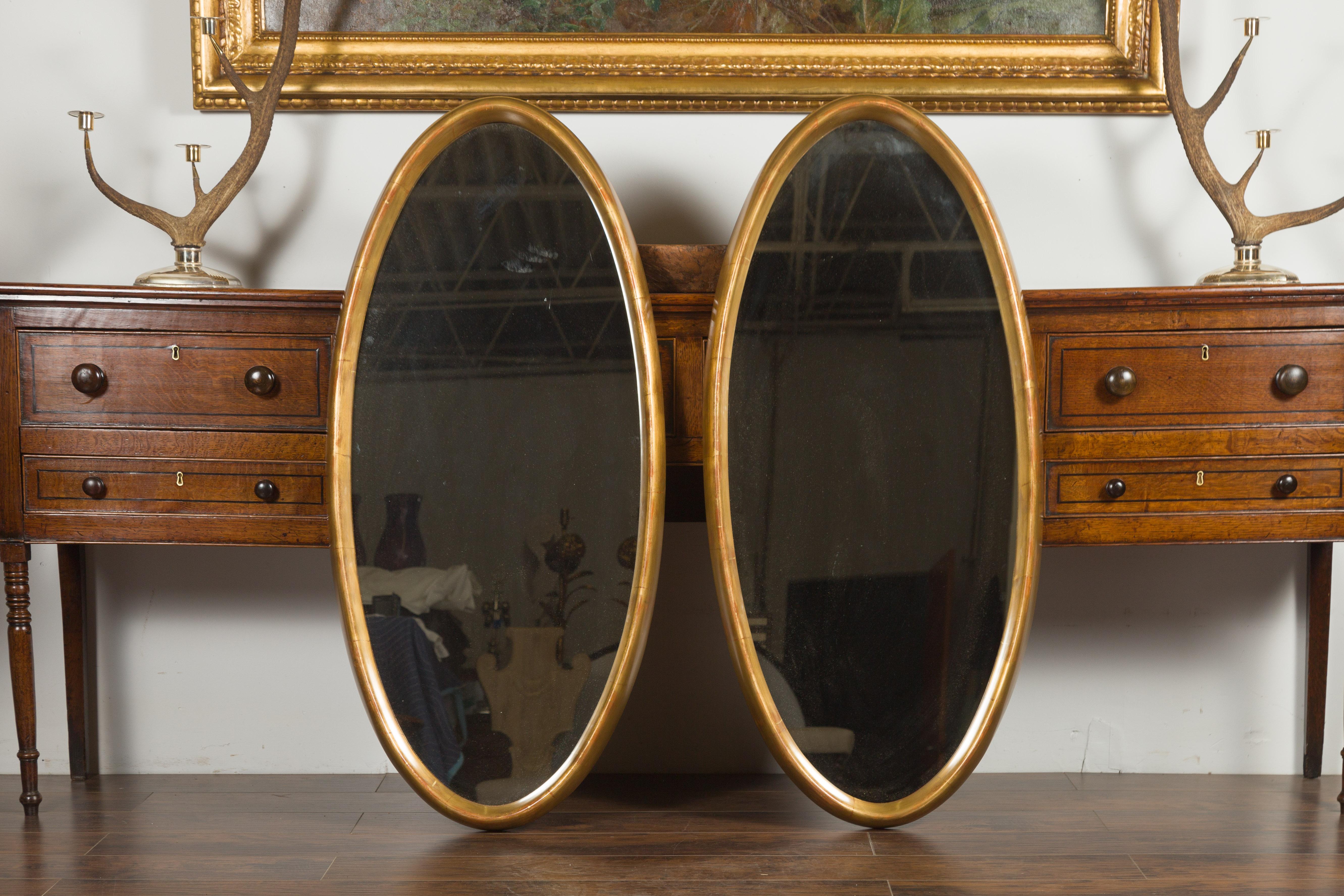 A pair of Italian vintage giltwood tall oval mirrors from the mid-20th century. Stylish and simple all in one breath, this pair of oval giltwood mirrors charms us with their clean lines and serene silhouette. Boasting a slight red undertone and each