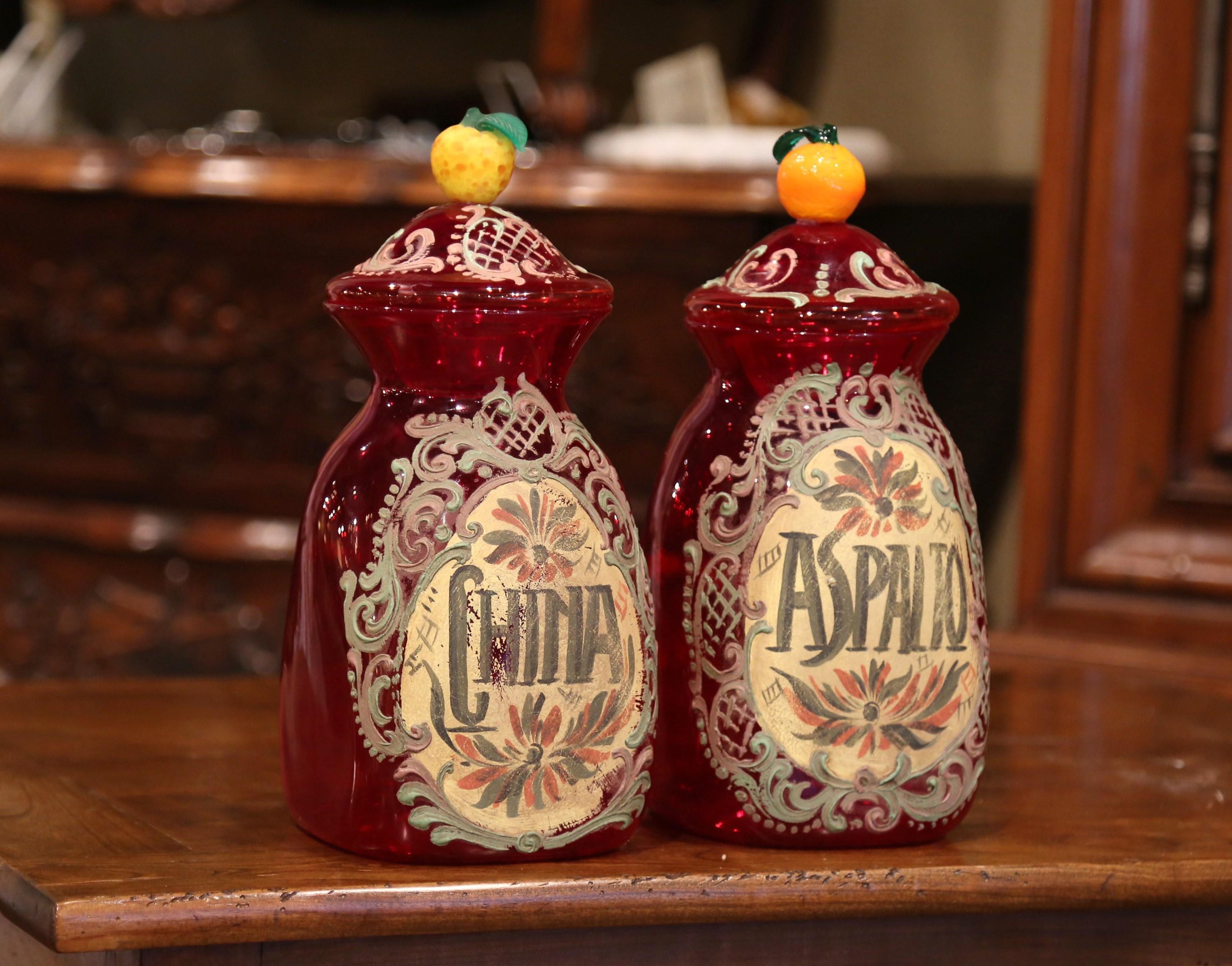 Decorate your bathroom with this pair of colorful antique painted apothecary jars. Crafted in Italy circa 1920, each colorful jar is made of red blown glass and each lid is embellished with a tangerine knob with leaf decor. The jar is decorated with