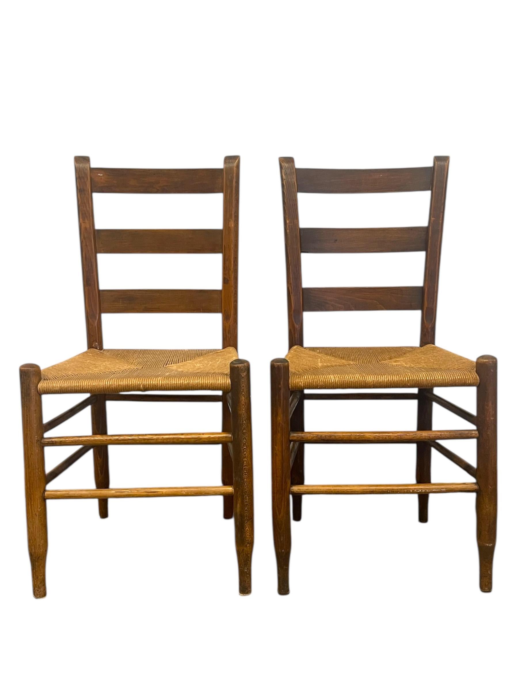   Pareja de sillas de comedor vintage con respaldo de escalera, asientos de junco tejido y armazón de madera. Estas sillas presentan respaldos de listones clásicos, camillas torneadas y patas cónicas que muestran una cálida pátina y un ligero