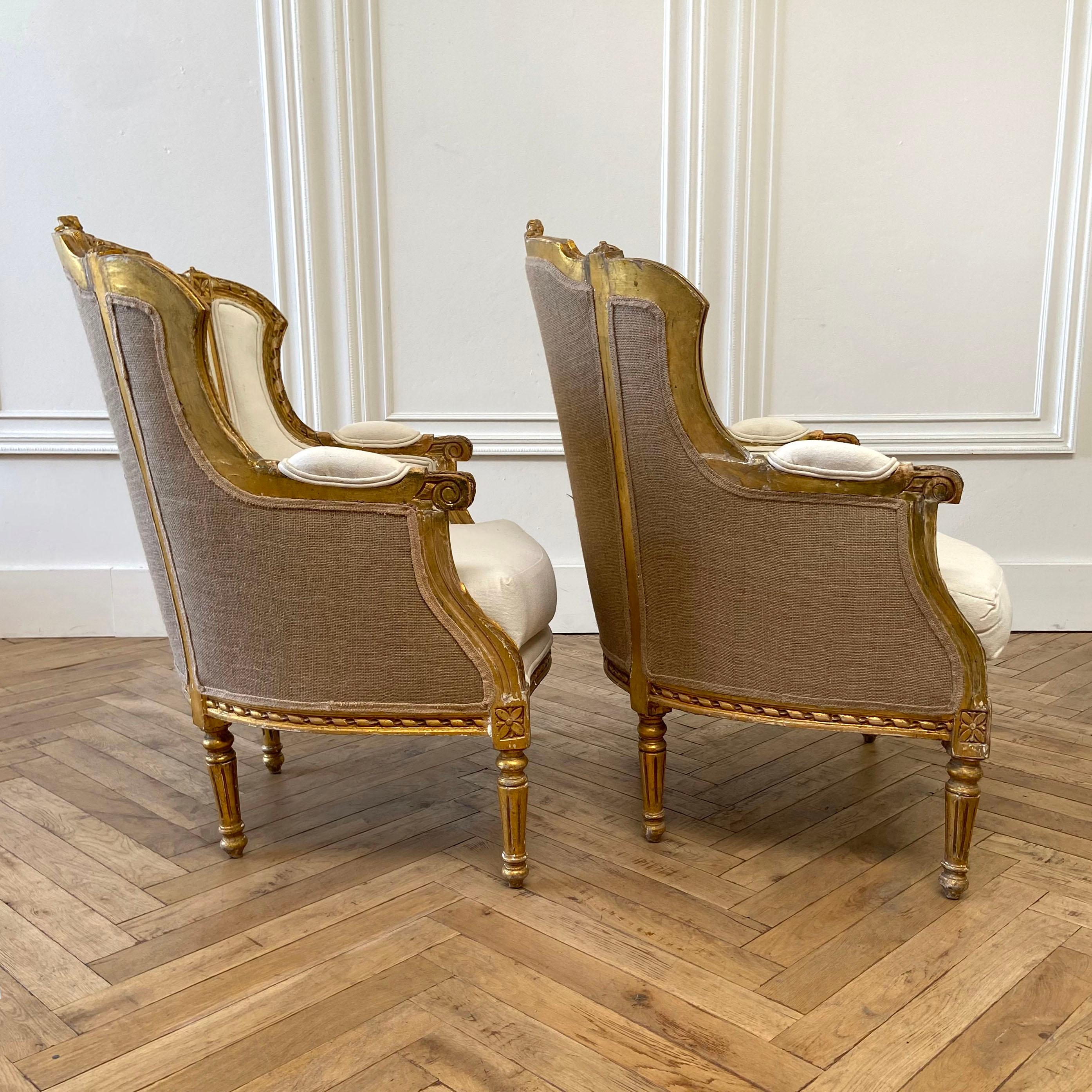 Pair of vintage Louis XVI style wing back bergère chairs upholstered in a natural canvas duck. Carved with large roses and ribbon detailing at the top of chairs and classic fluted legs, with a medallion carving. Natural distressed wood patina,