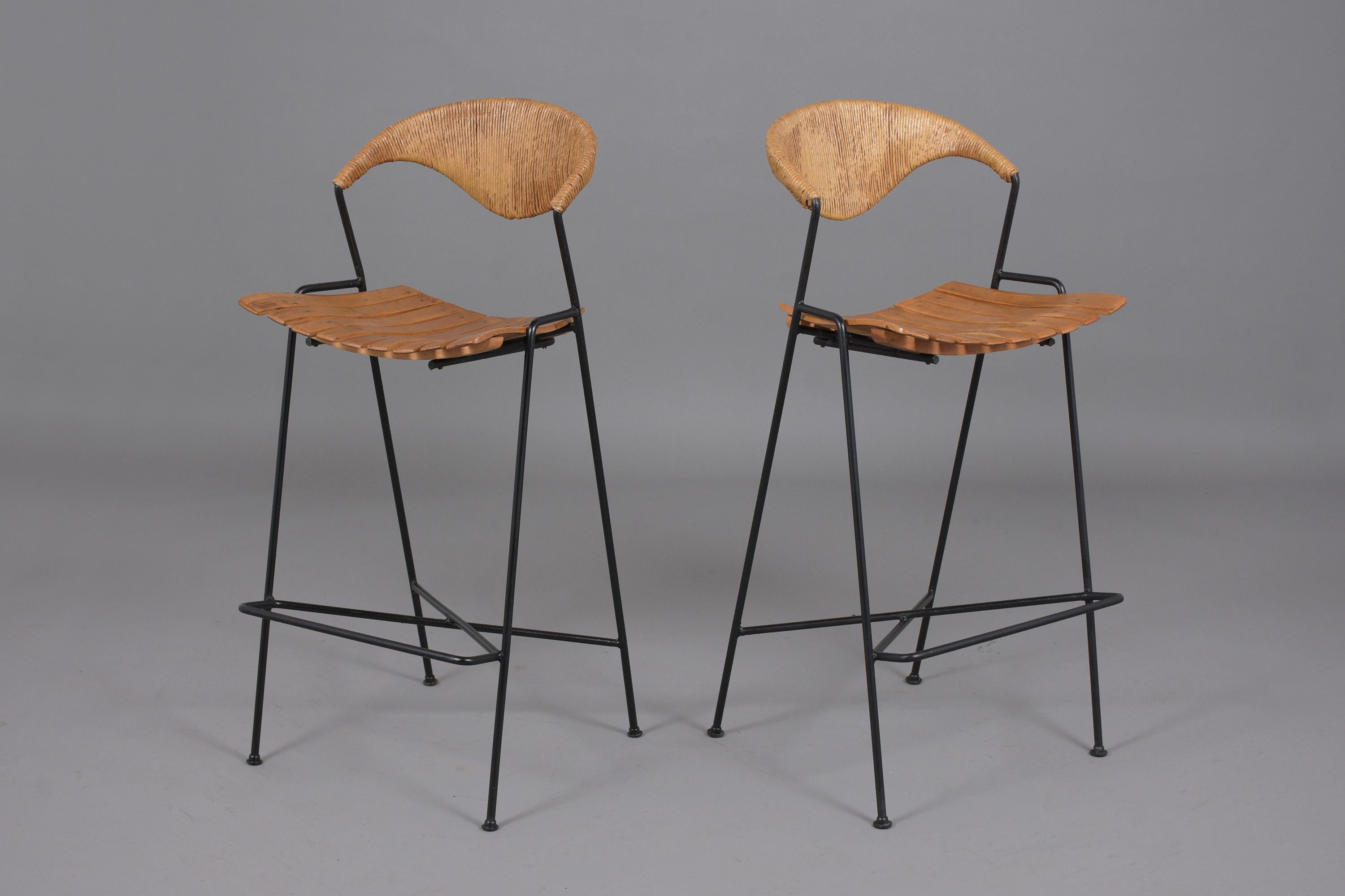 An excellent pair of Mid-Century Modern bar stools in the style of Arthur Umanoff that feature sturdy hand-crafted iron frames. This fabulous pair of barstools are in great condition, comes with backrests covered in a rush material, and wood panel