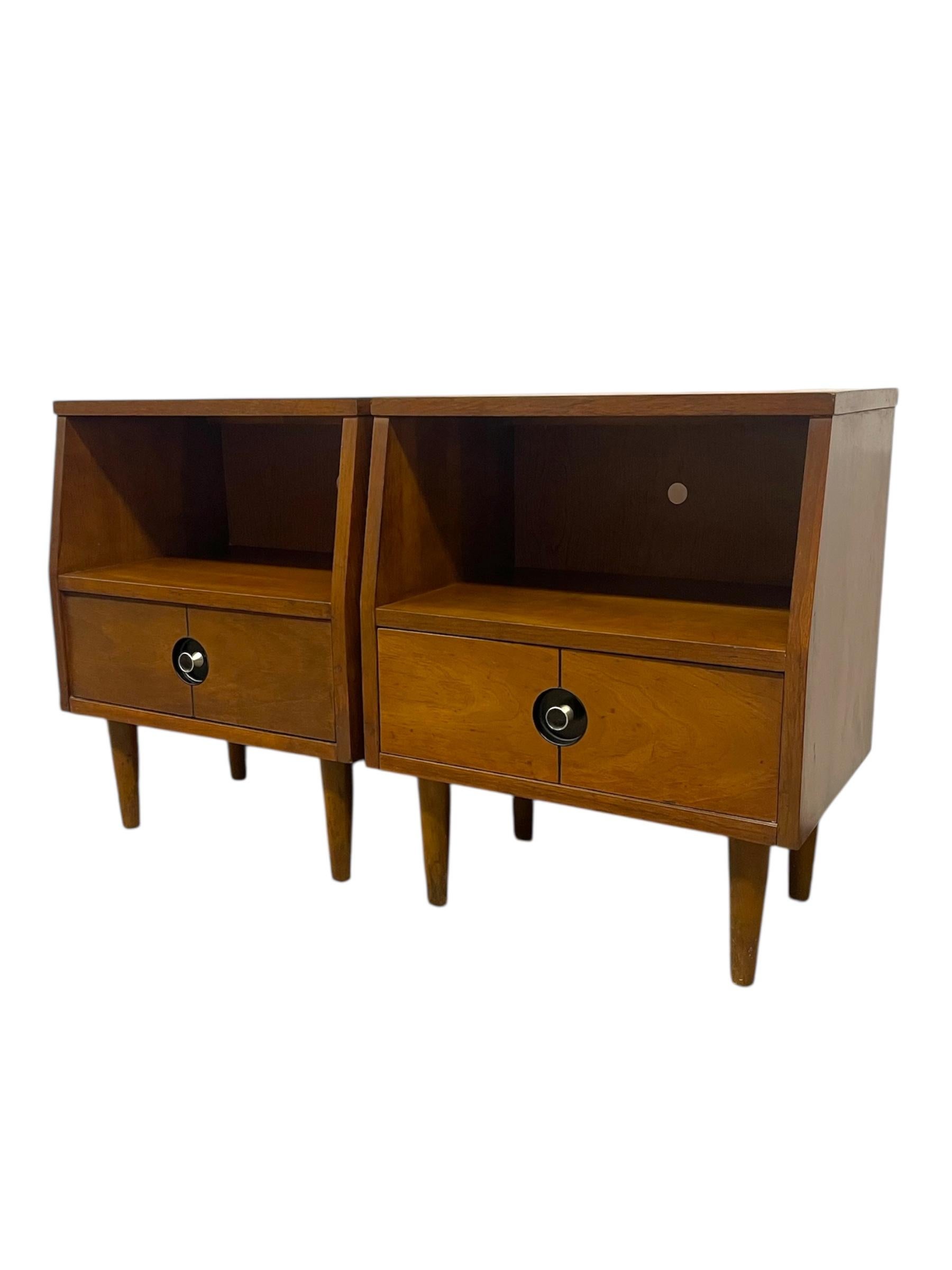  Matching pair of vintage mid-century modern nightstands with tapered legs and signature circular black-and-brass toned drawer pulls. Each unit features a single drawer below an open cubby, offering both concealed storage and easy-access