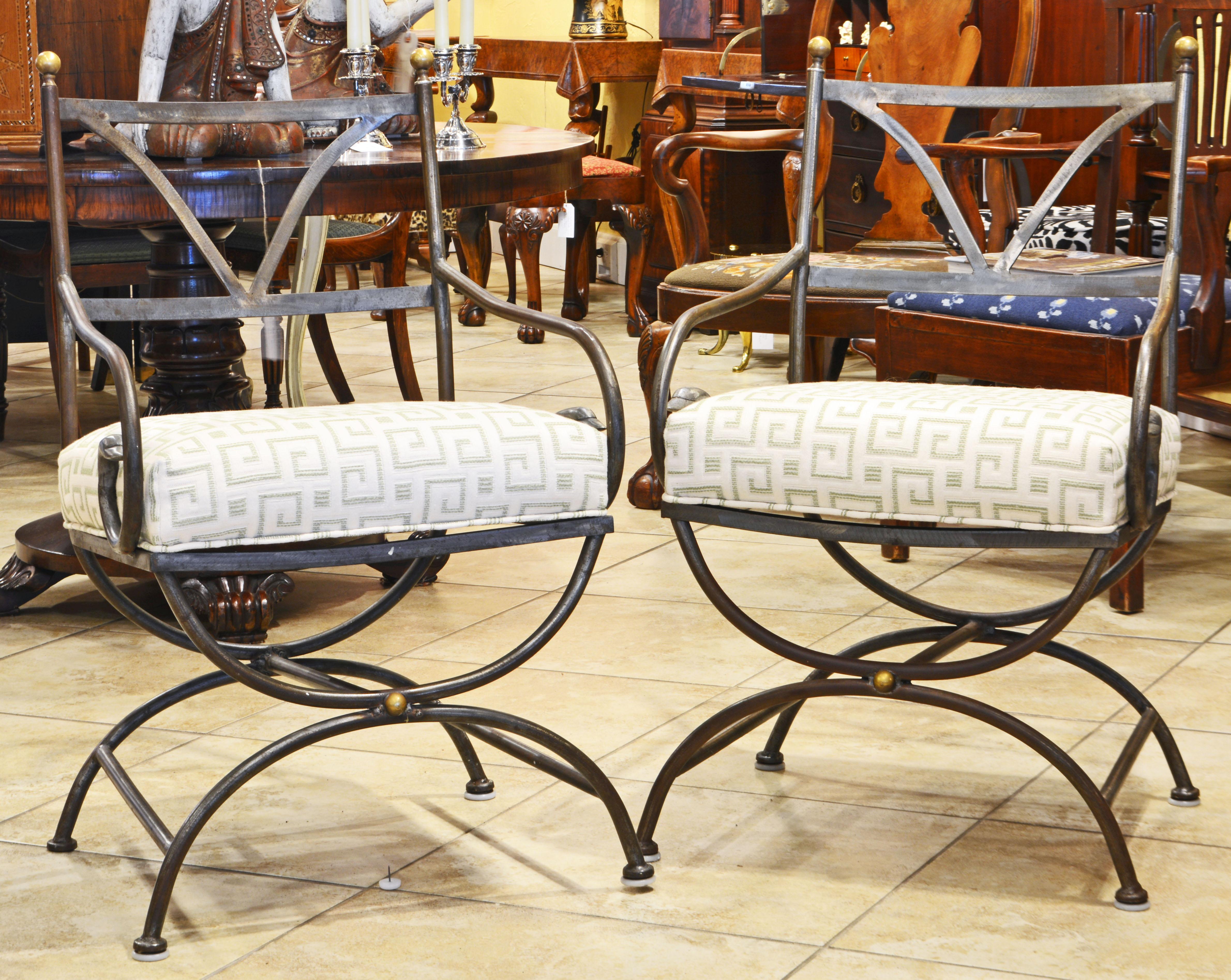 These wrought iron armchairs are designed in the classical curule style and feature curved armrests ending in stylized swan heads. The back rests are accented by mounted brass finials. The seats are upholstered and covered with a Greek Key themed