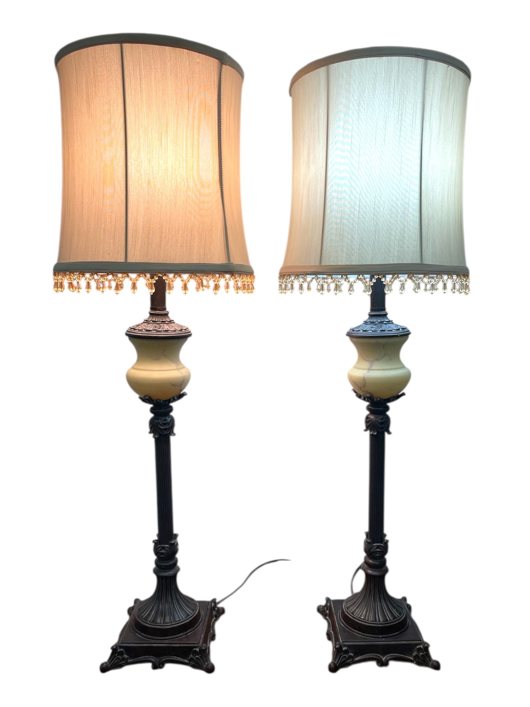 This pair of vintage table lamps features an elegant neoclassical-inspired silhouette with finely detailed cast-metal bases and columnar stems. Each lamp includes a fluted central column, decorative acanthus-style accents, and a square pedestal base