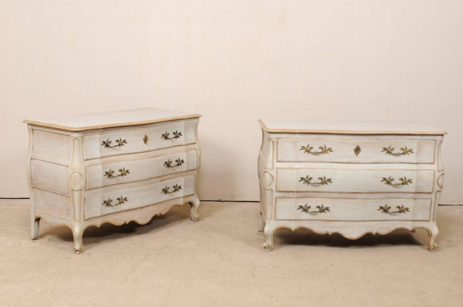 A pair of American vintage painted wood chest of drawers. This pair of American chests from the mid-20th century feature subtle curvy bombé style cases, scalloped skirt along three sides, decorated carvings down each side post, and paneled sides.