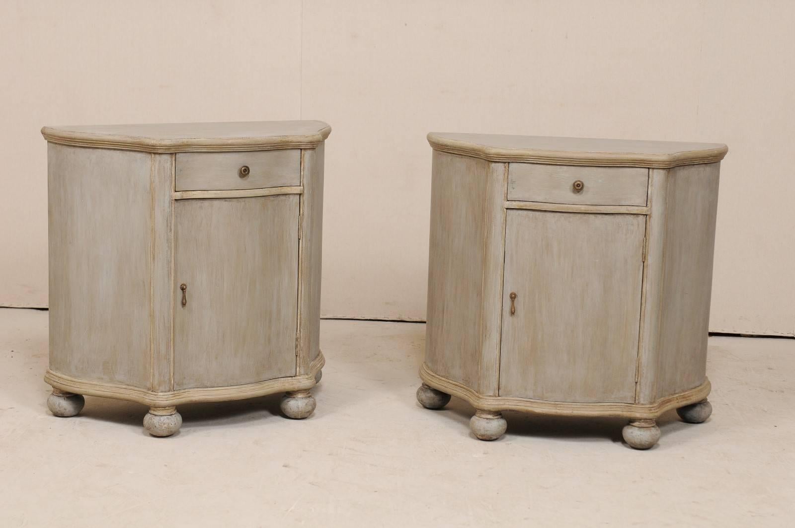 A pair of vintage American painted wood cabinets. These chests each feature demi styled bodies with curvaceous fronts and flat backsides. The cabinet body has a gentle undulating design along it's front and outward canting sides, giving these chests