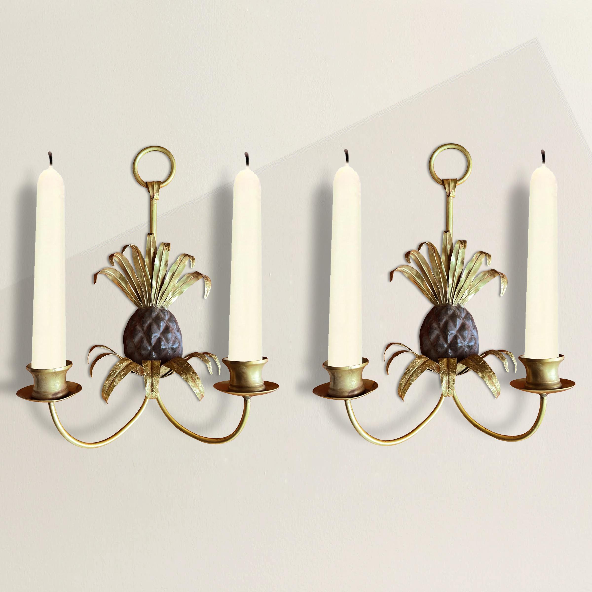 A chic and playful pair of Mid-20th Century French pineapple candle sconces with stamped brass crowns and leaves, arms and candle cups, and cast iron fruits. Pineapples have been symbols of friendship since the Colonial period in the US, and