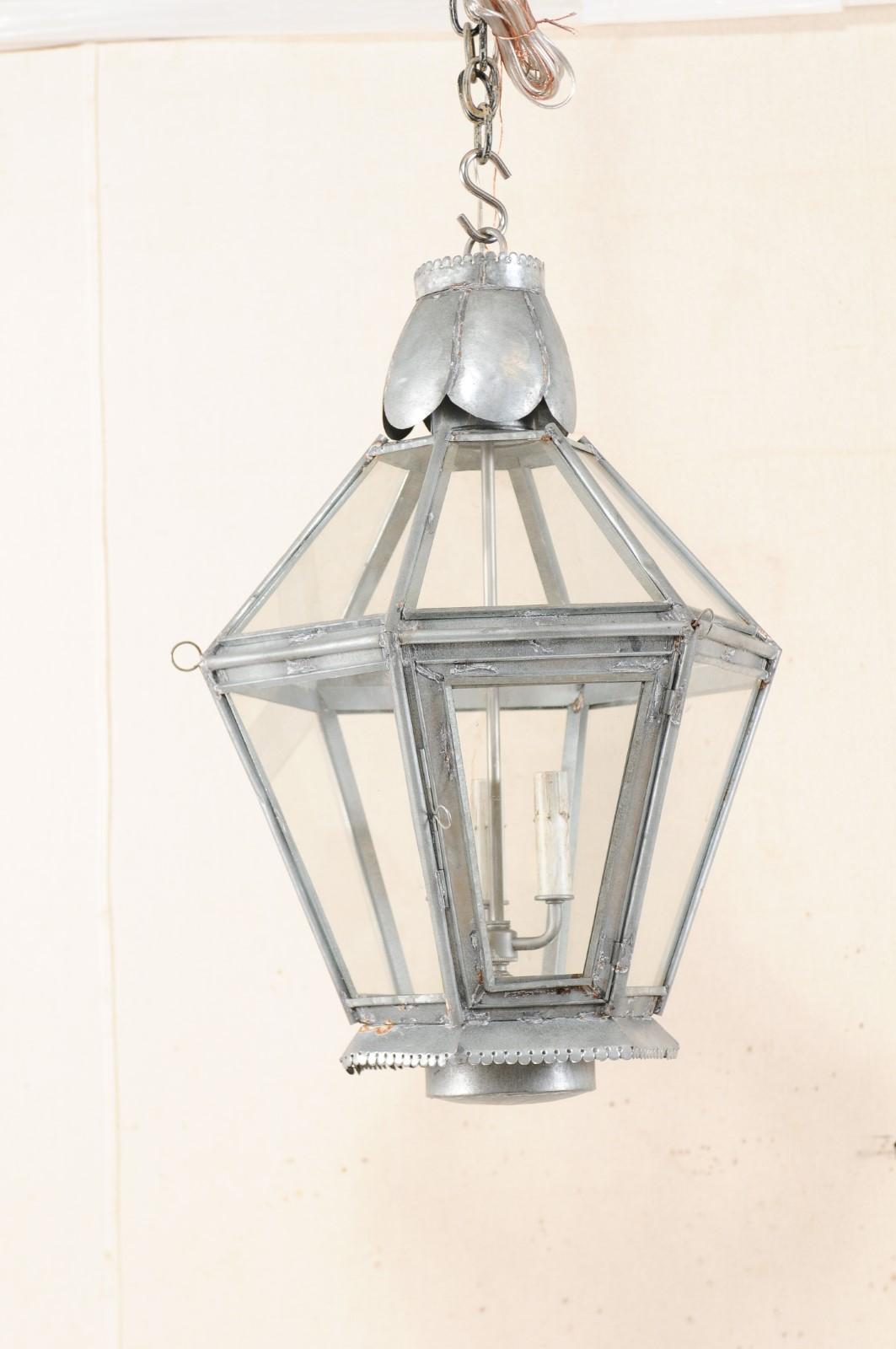 A pair of vintage hanging lanterns with three lights at center. Each lantern has a primarily hexagon-shaped body, with glass windows set within the silver metal frame which houses three lights topped with painted candle covers at their centers. A