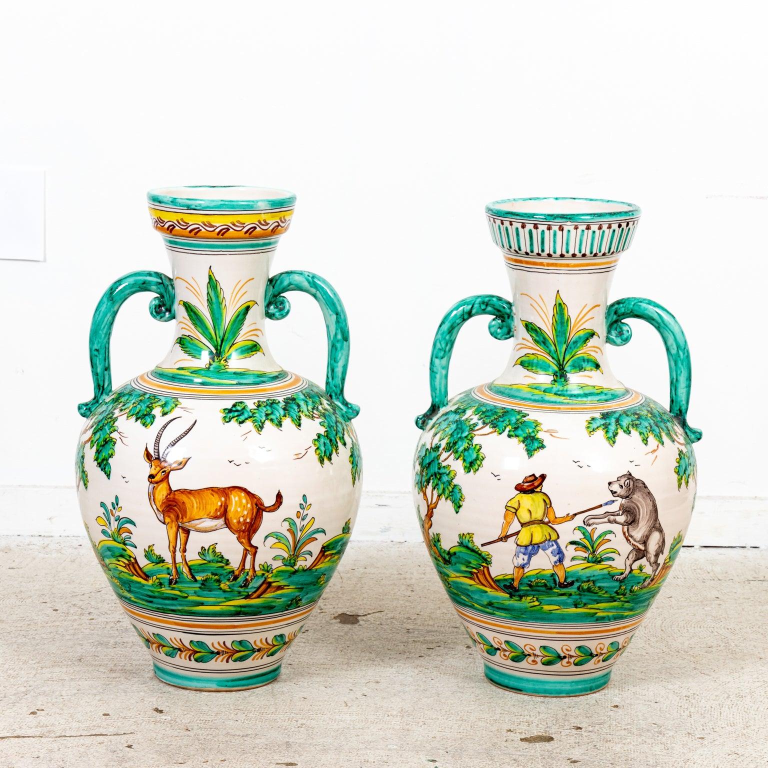 Pair of Spanish Majolica urns in a glazed finish with double handles and floral painted trim. The body of the urns are detailed with a deer illustration, a depiction of a man in an attack stance with a spear fighting against a bear, and a large