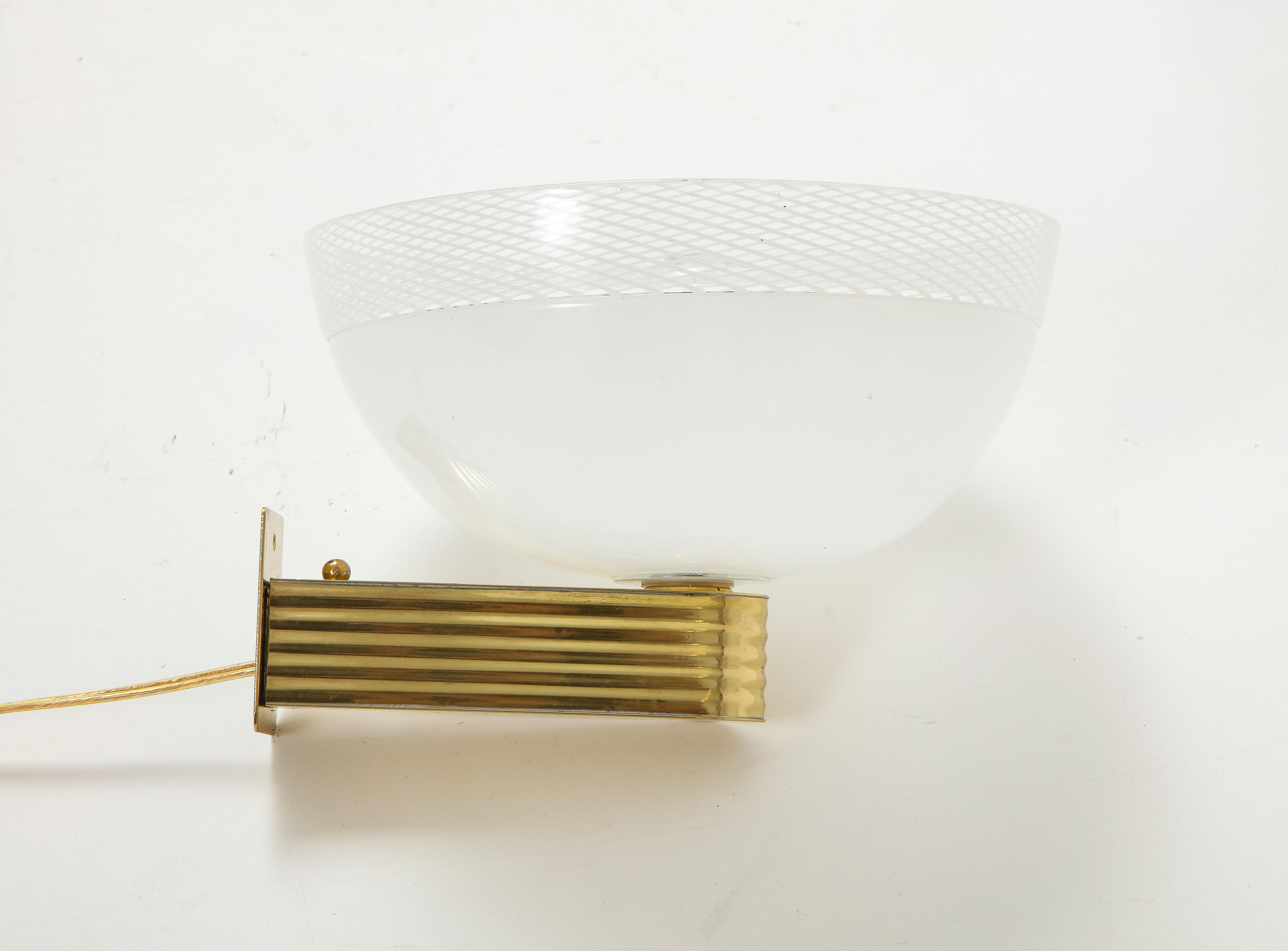An elegant and rare pair of Venini Murano glass wall lights; the delicate opaline circular glass light is complemented at the top border with the precious reticello technique. The glass is supported by a ribbed brass base and wall support.
