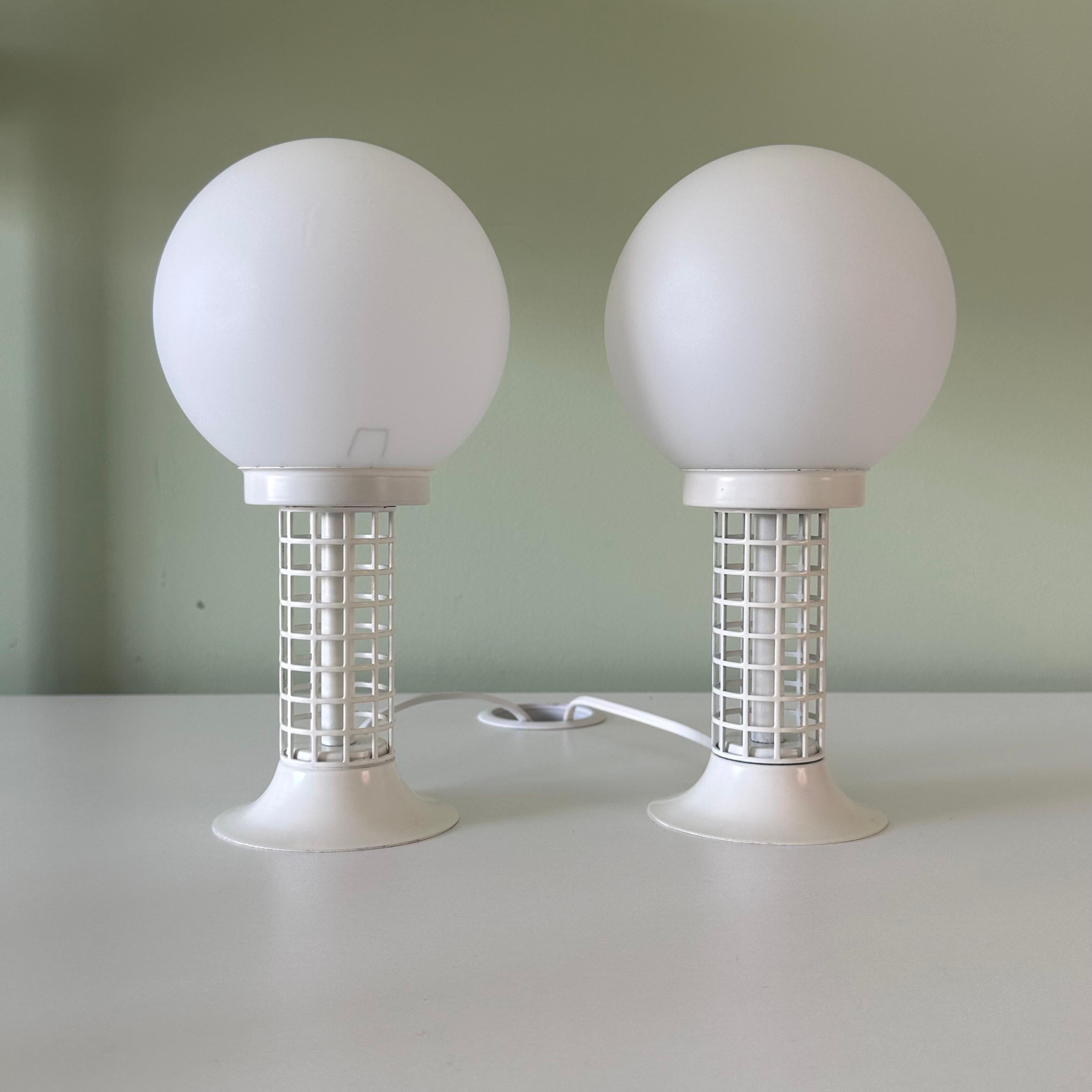 Pair of vintage 1970s modernist style vintage table lamps. Perfect all white look. The top half features white frosted spherical glass globe shades, supported by a white painted metal base with windowpane, grid or perforated motif. Socket mounted