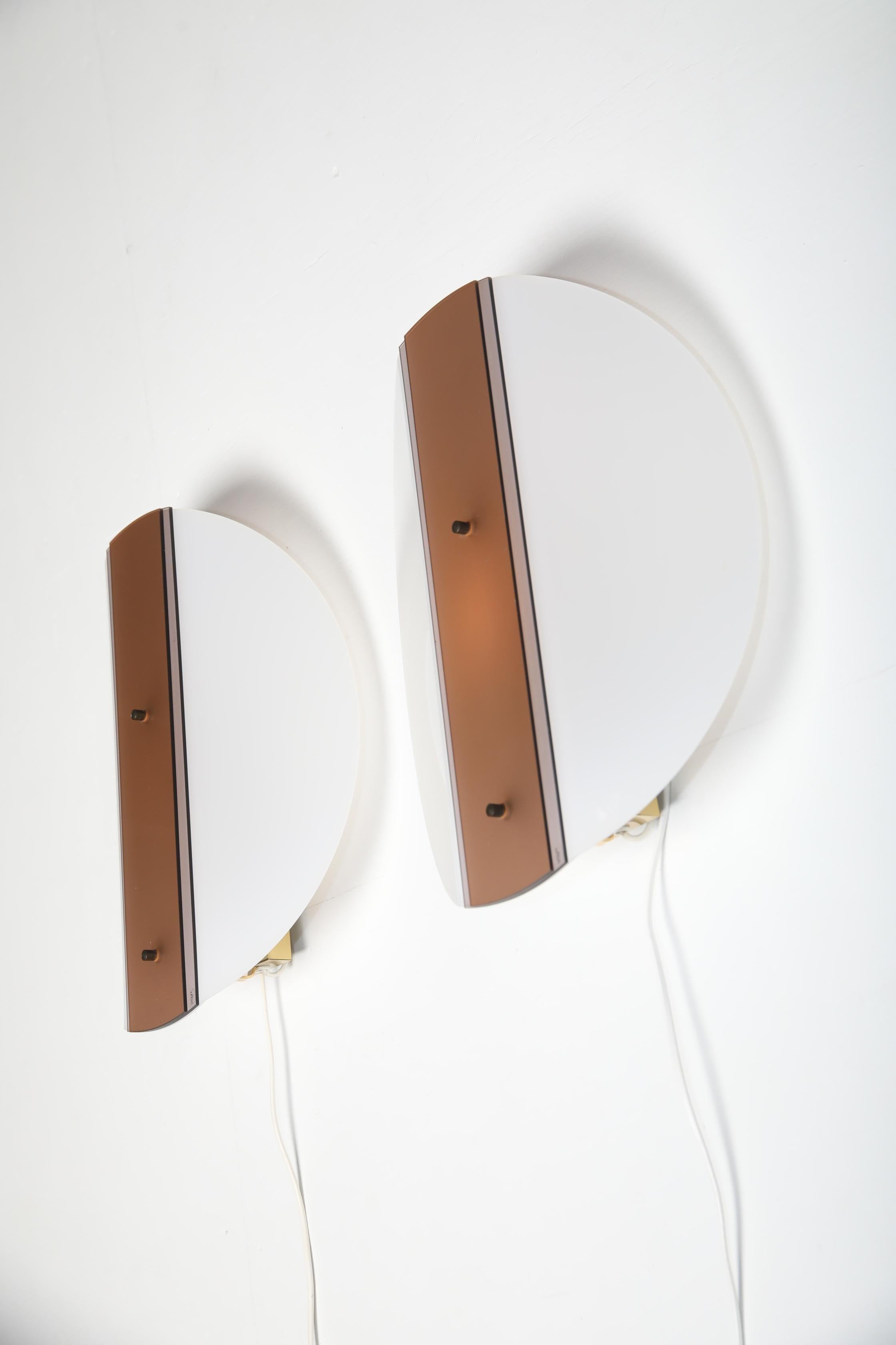 Pair of wall lamps produced by Artlight Design in the 1980s. Each lamp is made of plastic and metal and consists of a circular front panel combined with a vertical centre section that holds the switch and mounting parts. The lamps have a clear,