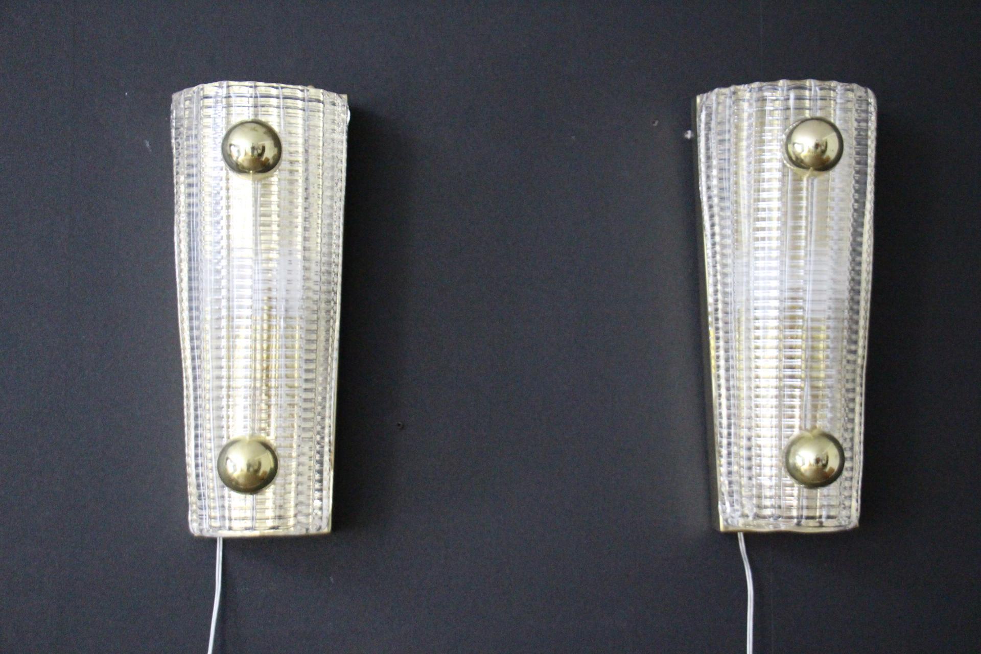 Cette magnifique paire d'appliques a été fabriquée à Murano . Elles présentent un aspect classique et très moderne grâce à leur beau verre de cristal texturé. Leur forme est très stricte et s'adoucit grâce à ses boules en laiton doré.
Elles reposent