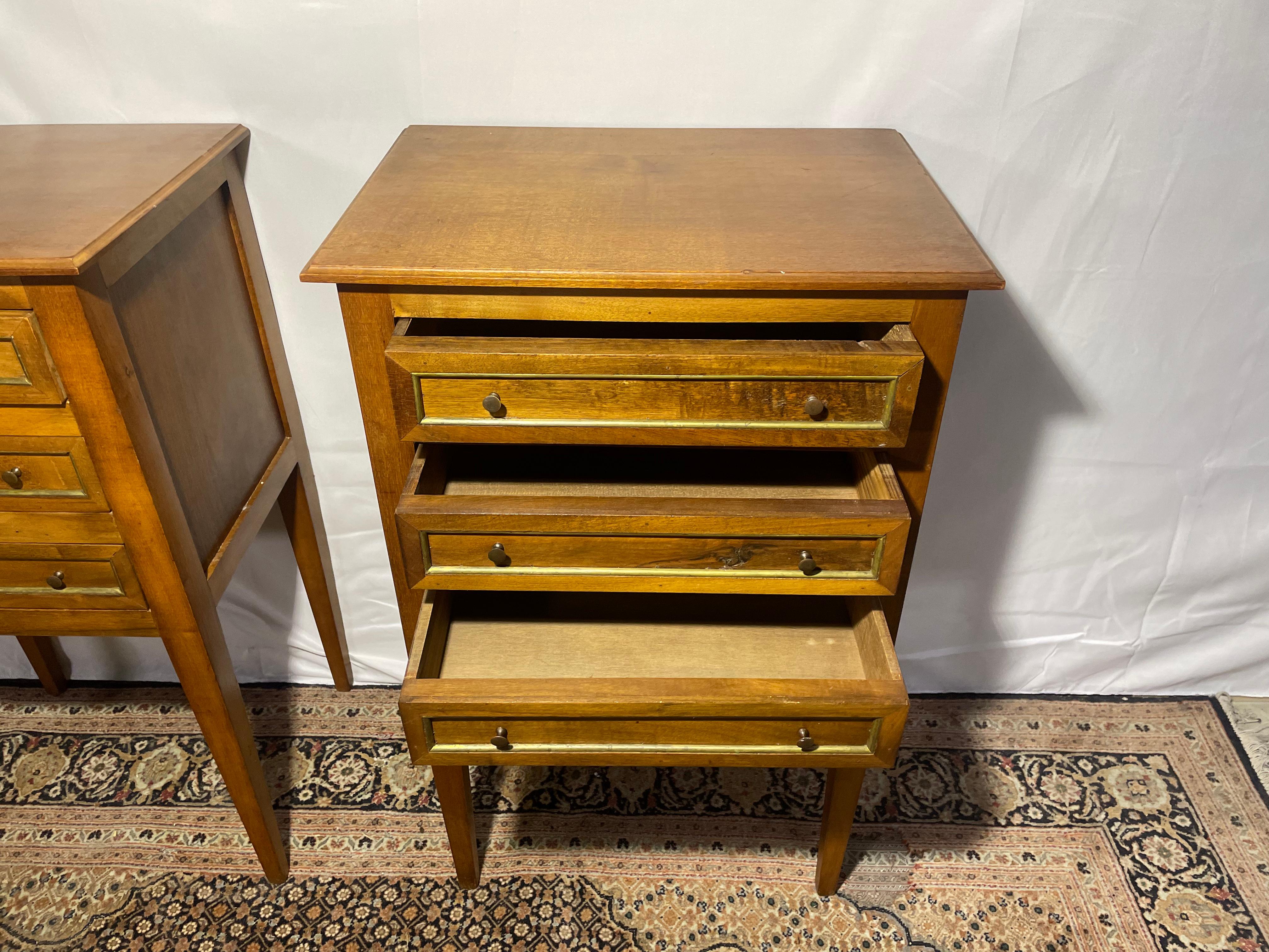 This elegant pair of early 20th-century nightstands is crafted from solid walnut, featuring a refined Louis XVI-inspired design. Characterized by tapered legs, three spacious drawers, and gold detailing, these pieces showcase the rich, warm tones of