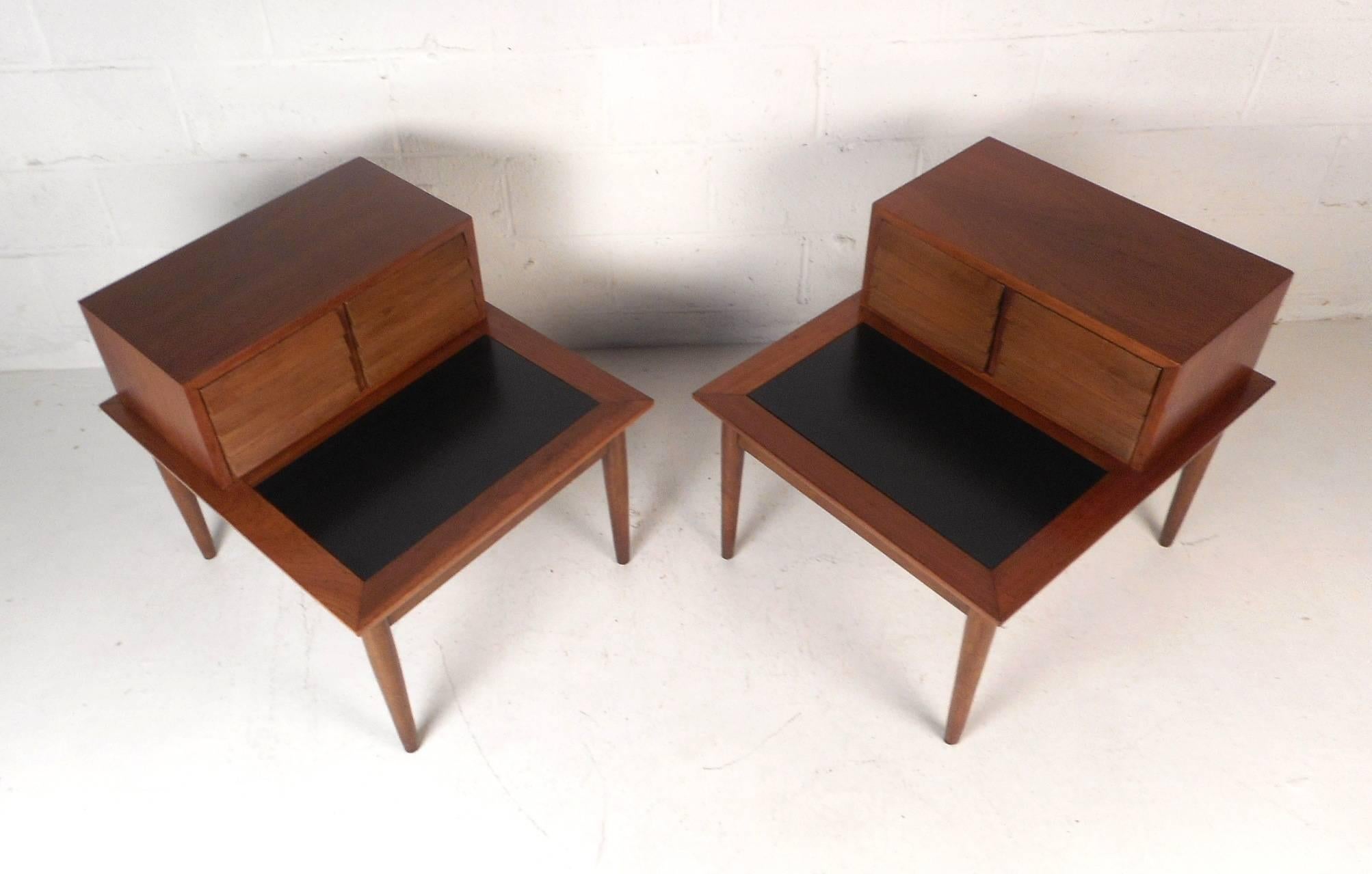 This stunning pair of vintage modern end tables feature two small louvered drawers and a bottom tier with a black laminate top. A sleek two-tier design made of walnut that sits on top of four tapered legs. Quality construction with four sculpted