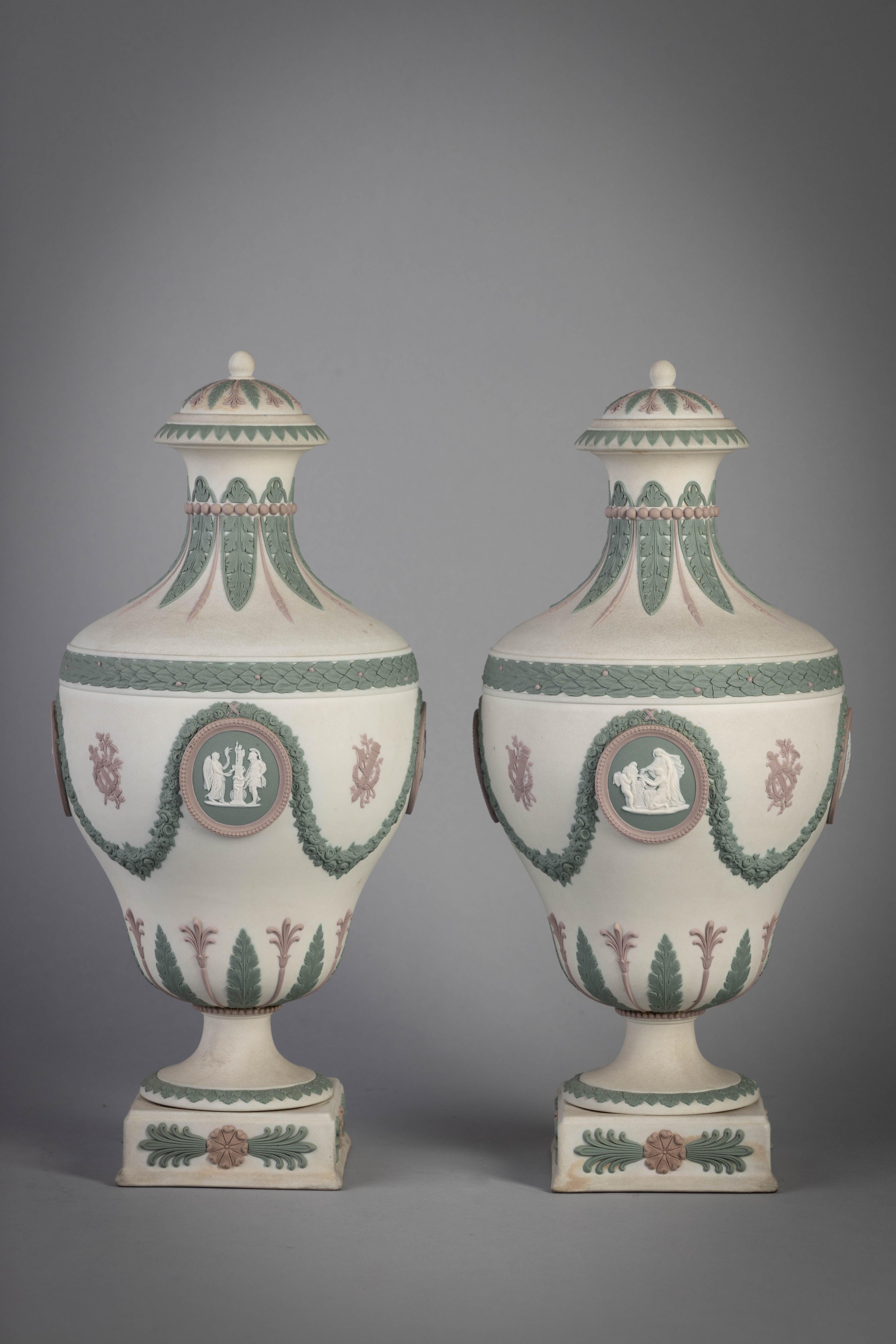 Sage green and lilac jasperware. Each pyriform vessel decorated with figural medallions beneath floral garlands, further decorated with leaf and palmette bands, the domed cover with ball finial on circular foot and square plinth, impressed WEDGWOOD,
