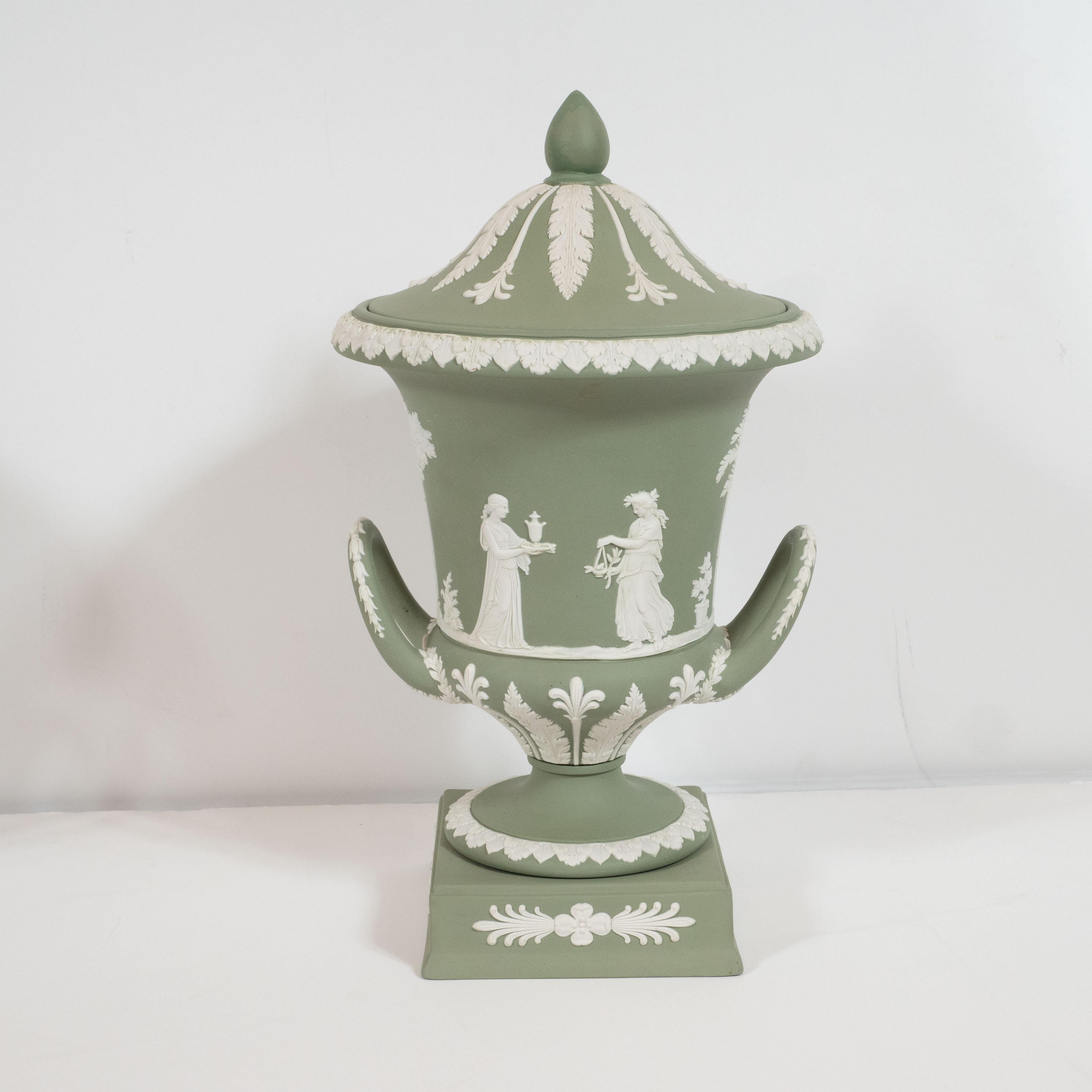 This elegant pair of neoclassical jasperware ceramic urns was realized by the esteemed British maker Wedgwood, circa 1950. They feature volumetric rectangular bases with a stepped bottom and a beveled top adorned on each side with raised