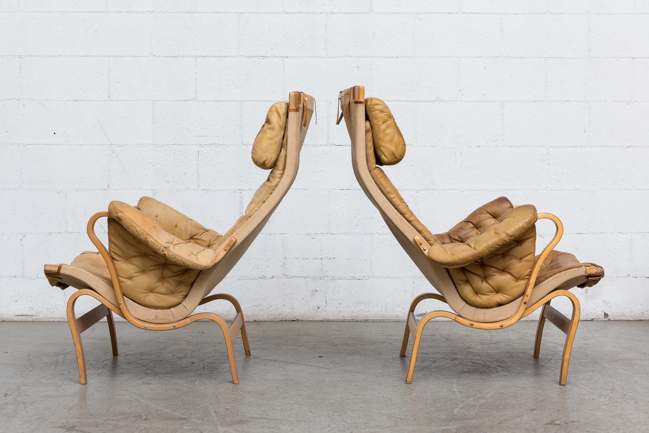 Very worn and well used pair of 1970s 'Pernilla' lounge chairs with arms by Bruno Mathsson for DUX. Completely original with extreme wear, one has visible bite marks from a dog. Natural leather and canvas. Sold as is, set price.
