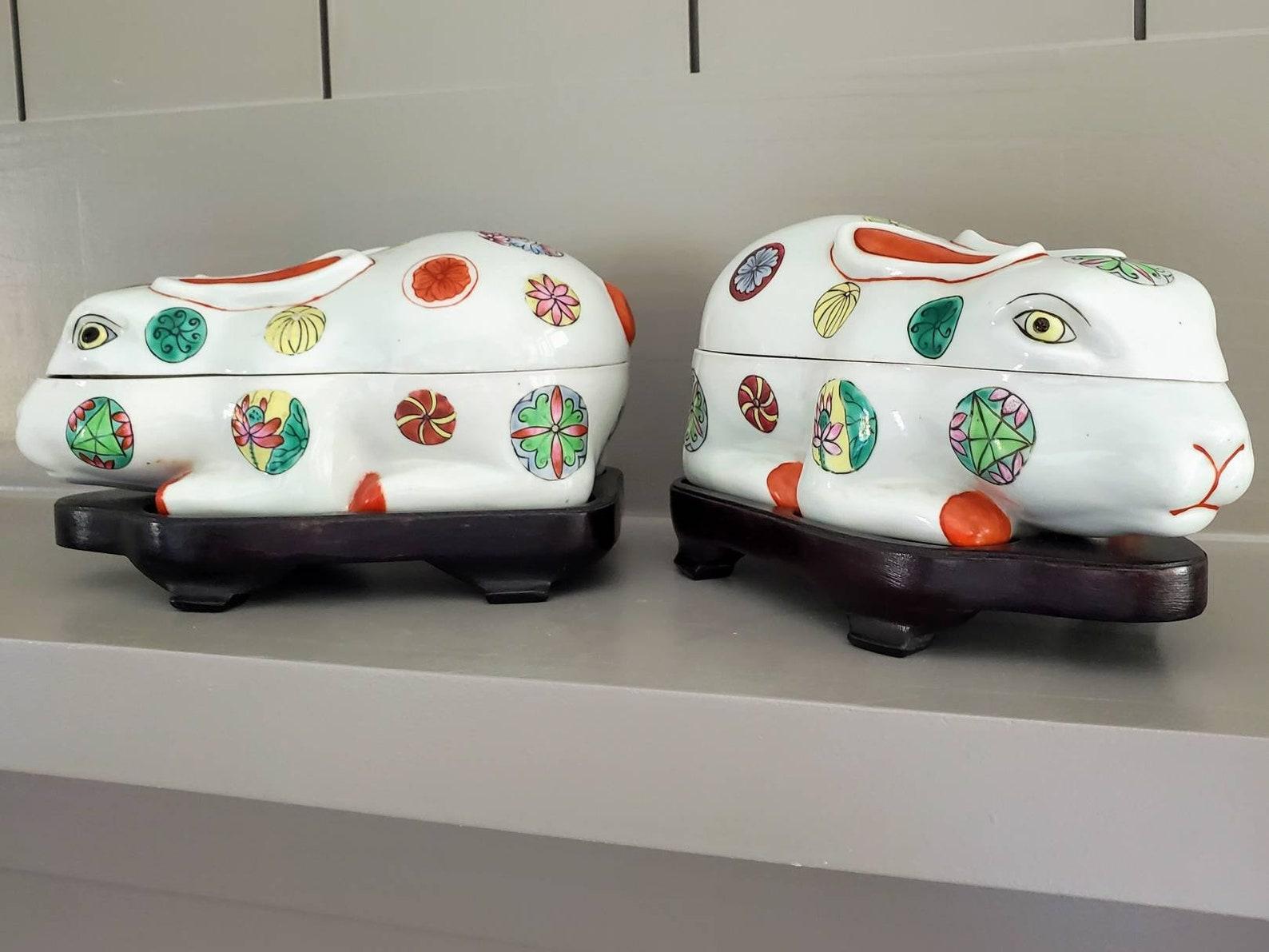 A fascinating pair of Japanese porcelain kogo (incense containers), crouching rabbit figures, hand painted, polychrome decorated with mon inspired symbols. The top half lifts off to reveal a spacious open interior. Resting on fitted wooden stands.