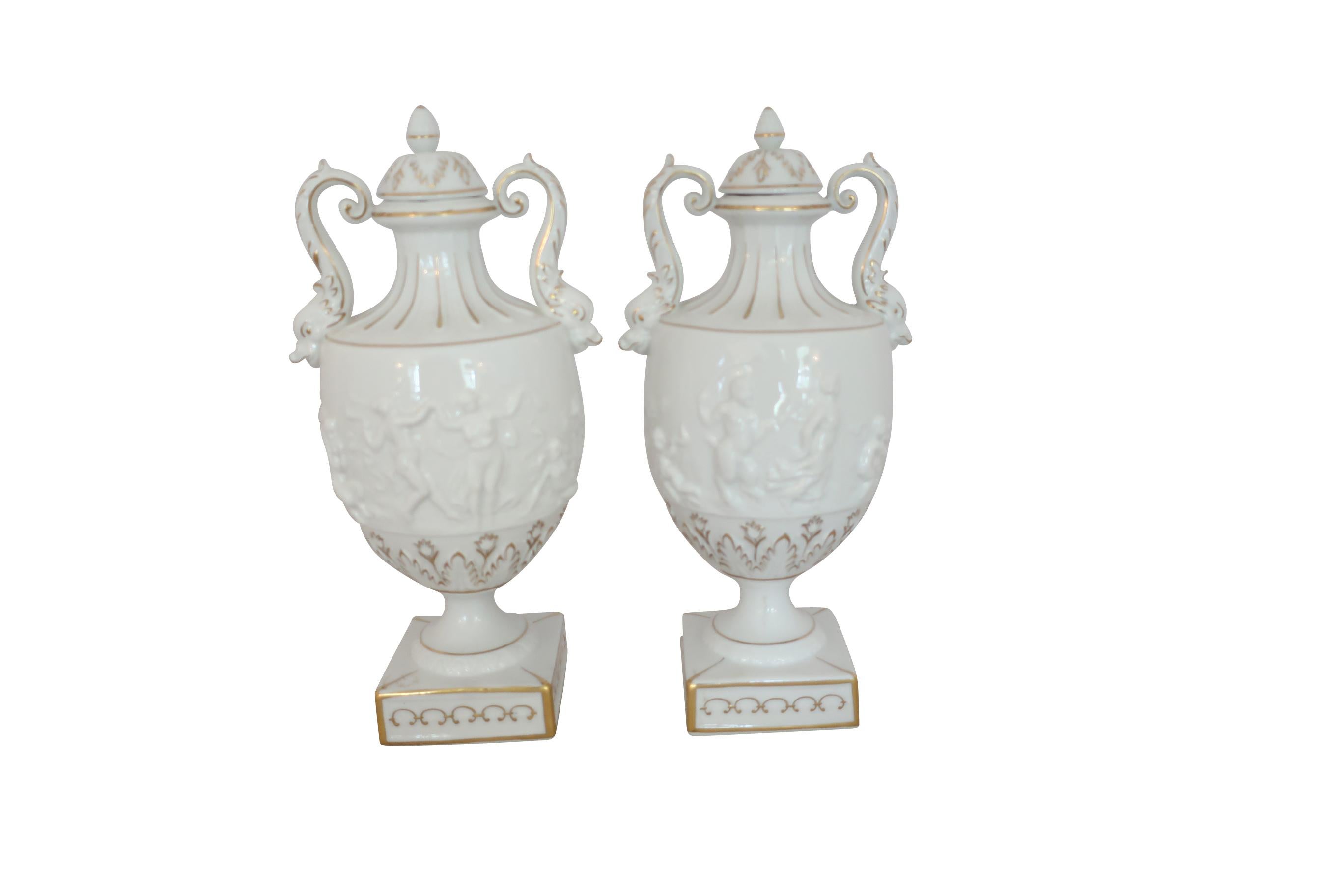 Pair of white and gilt hand painted Capodimonte classical urns with lids on a tapering square base. The urns have Classic scrolled handles and are decorated on the bodies with raised putti motifs. Small tulip-shaped gilt floral decorations below the
